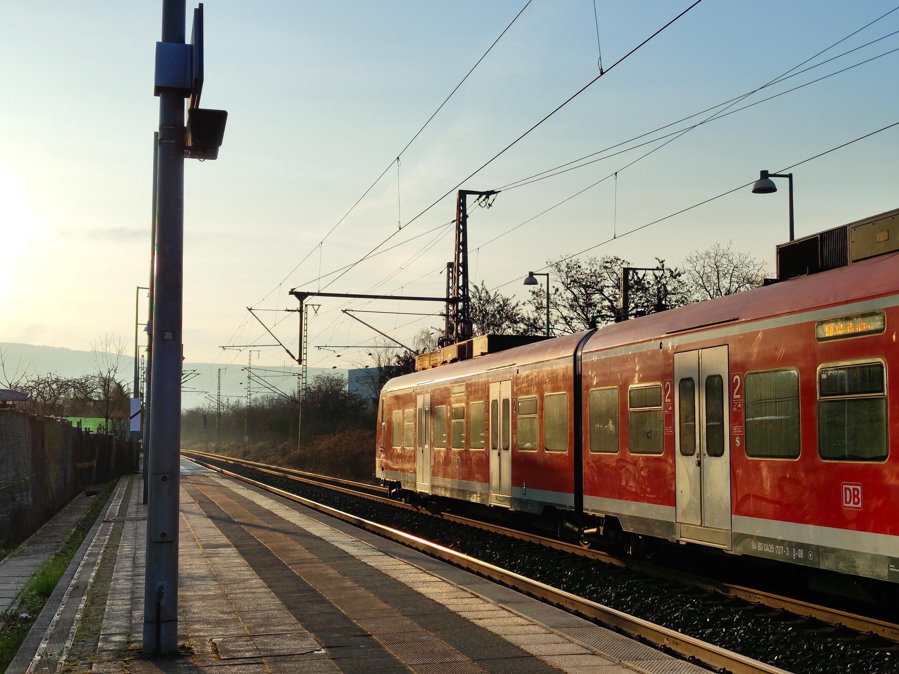 A red Deutsche Bahn commuter train just setting off on a railway track beside a platform under clear dawn skies.