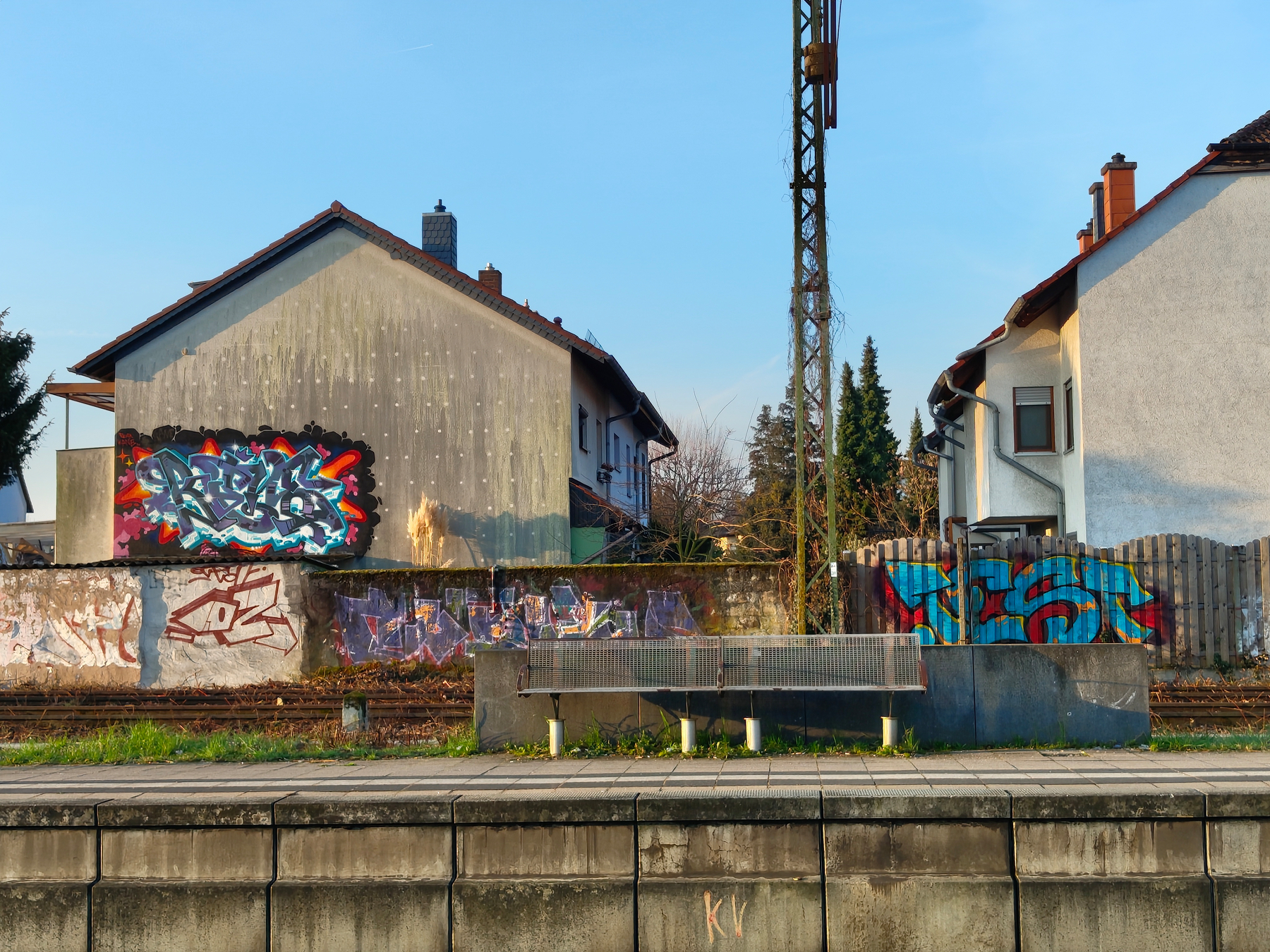 Two houses, one with a graffiti-covered wall, the other with graffiti on the fence, are seen behind a quiet railway platform and an empty bench.