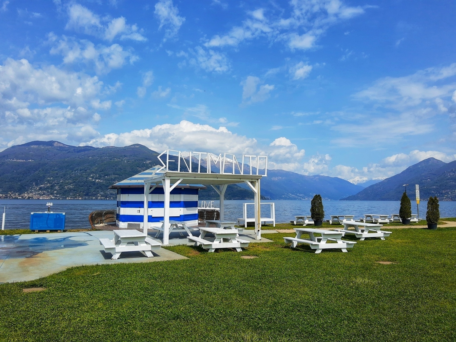 Chiringuito in riva al Lago Maggiore, sopra il locale si vede la scritta Sentimento. Sullo sfondo sono visibili il lago e le montagne.