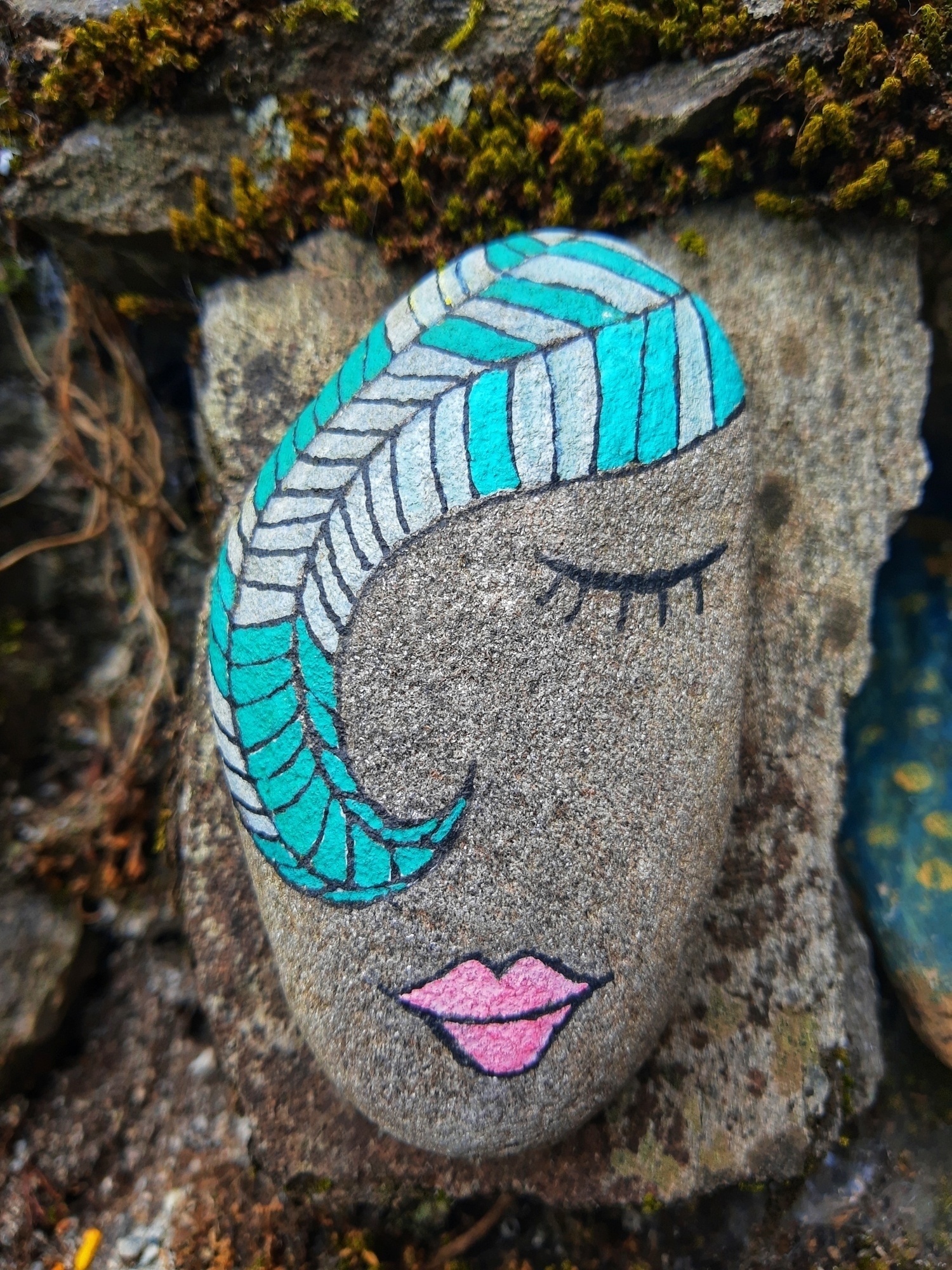 Auto-generated description: A painted rock features a face with closed eyes, pink lips, and turquoise and white striped hair, resting among natural stones.