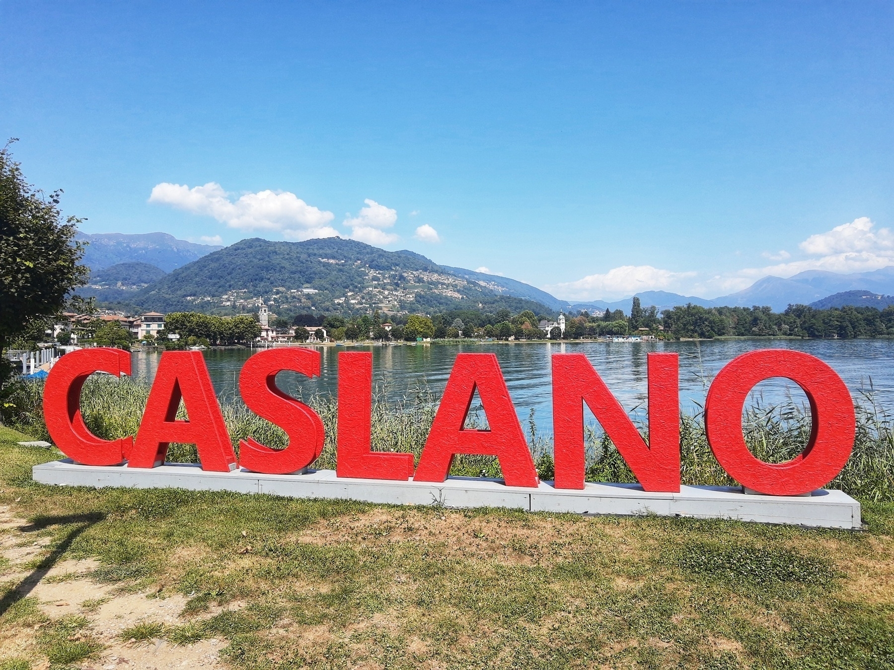 Auto-generated description: Red letters spelling CASLANO stand on a grassy area by a lake, with mountains in the background.