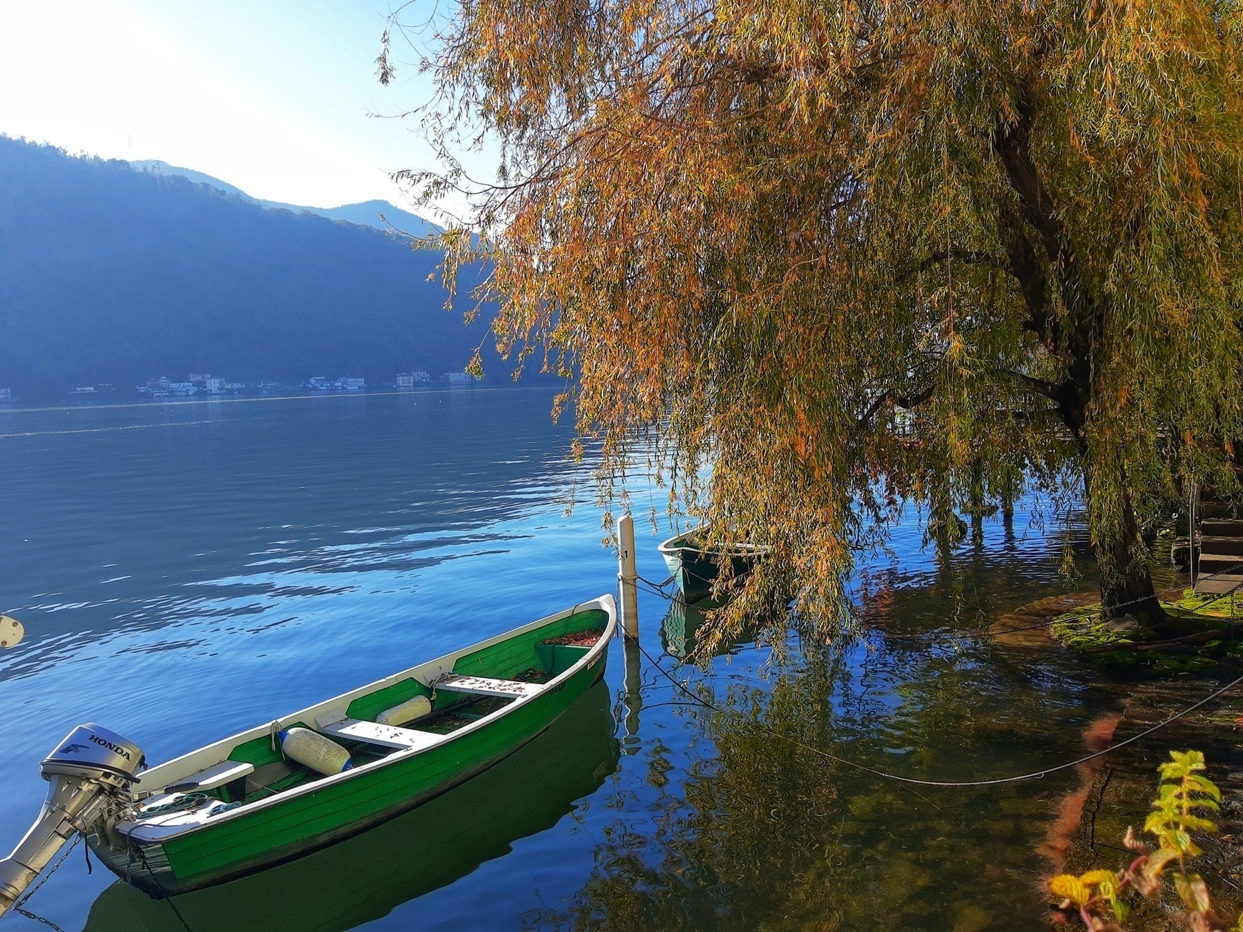 Auto-generated description: A green boat is docked beside a calm lake surrounded by autumn trees.