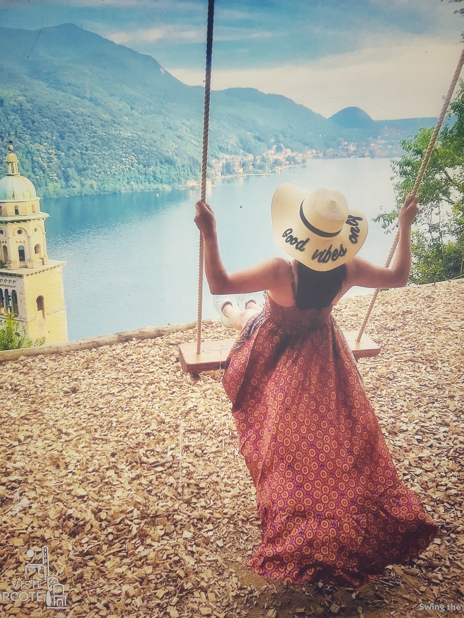 Auto-generated description: A person in a wide-brimmed hat and long dress swings over a scenic lake view with mountains in the background.