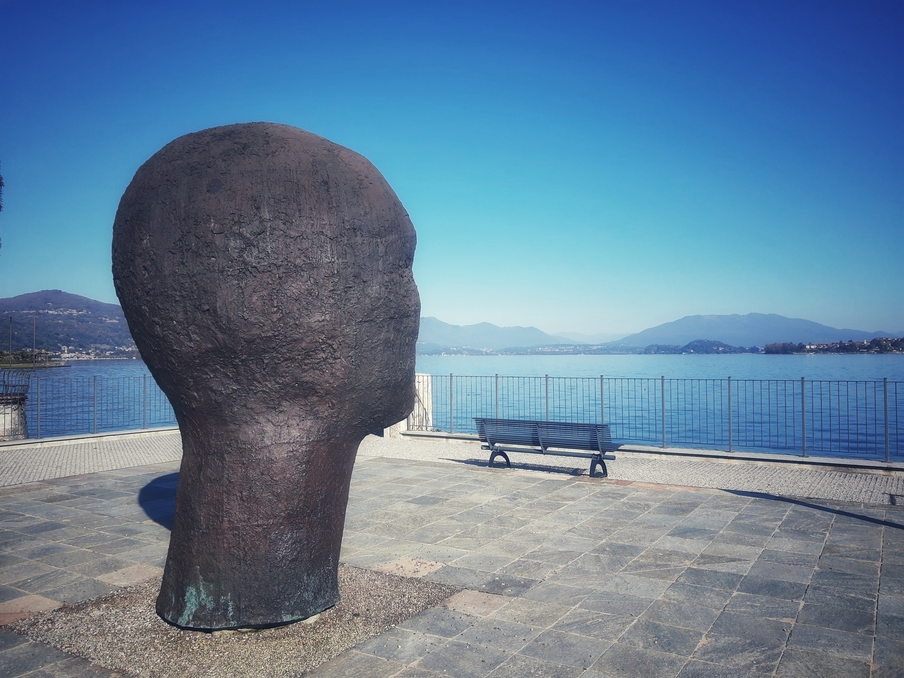 Auto-generated description: A large head sculpture overlooks a scenic lake with mountains in the background.