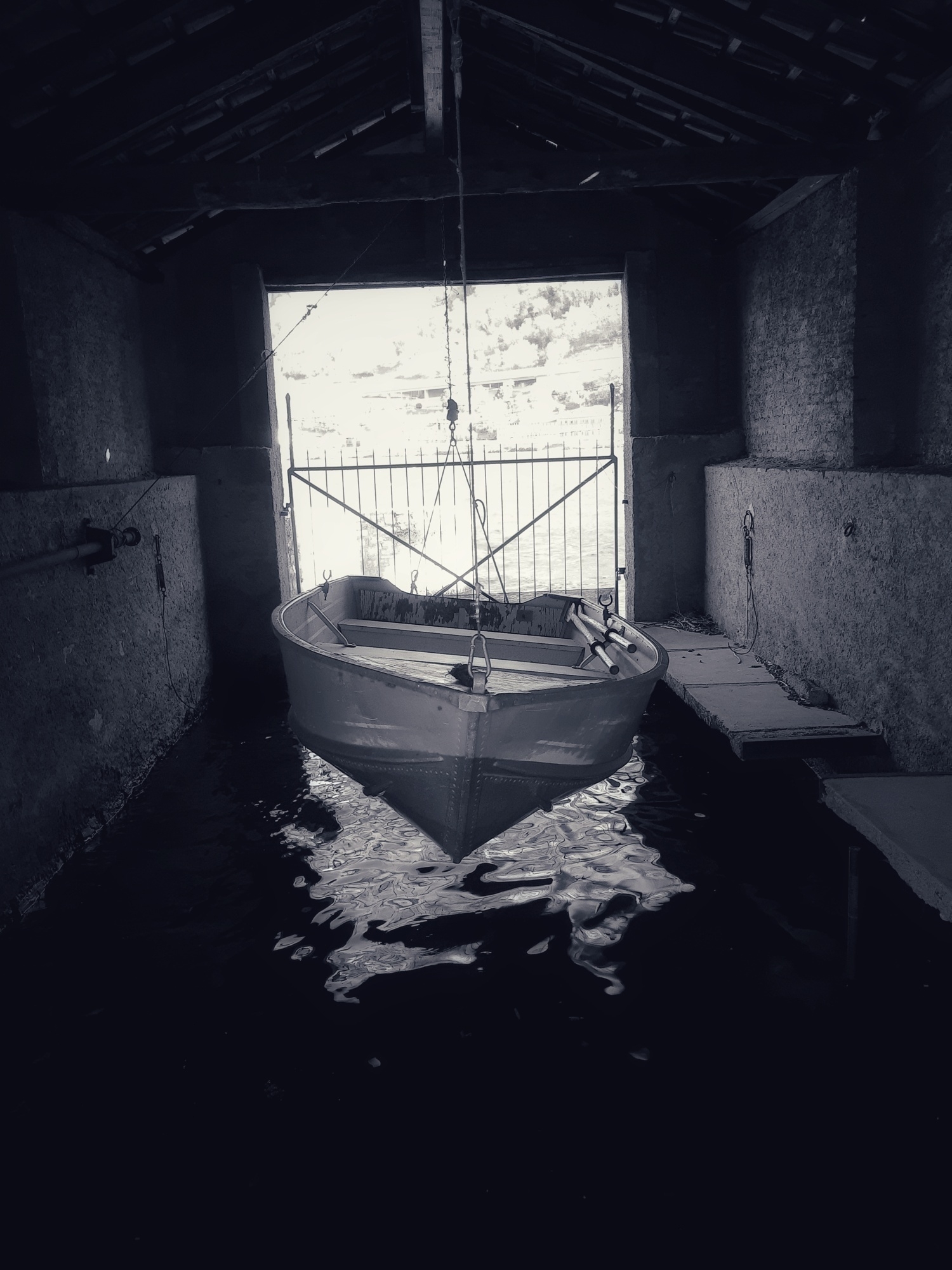 Auto-generated description: A small boat is suspended over water inside a dimly lit boathouse with an open gate.