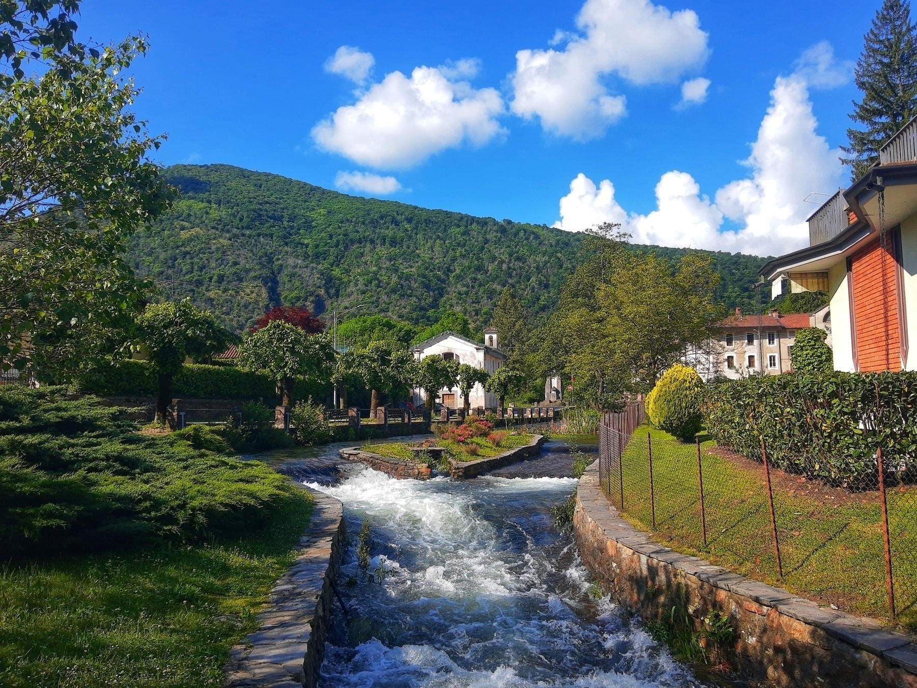 Auto-generated description: A serene landscape features a flowing river, lush greenery, and charming houses set against a mountainous backdrop with a blue sky and fluffy clouds.