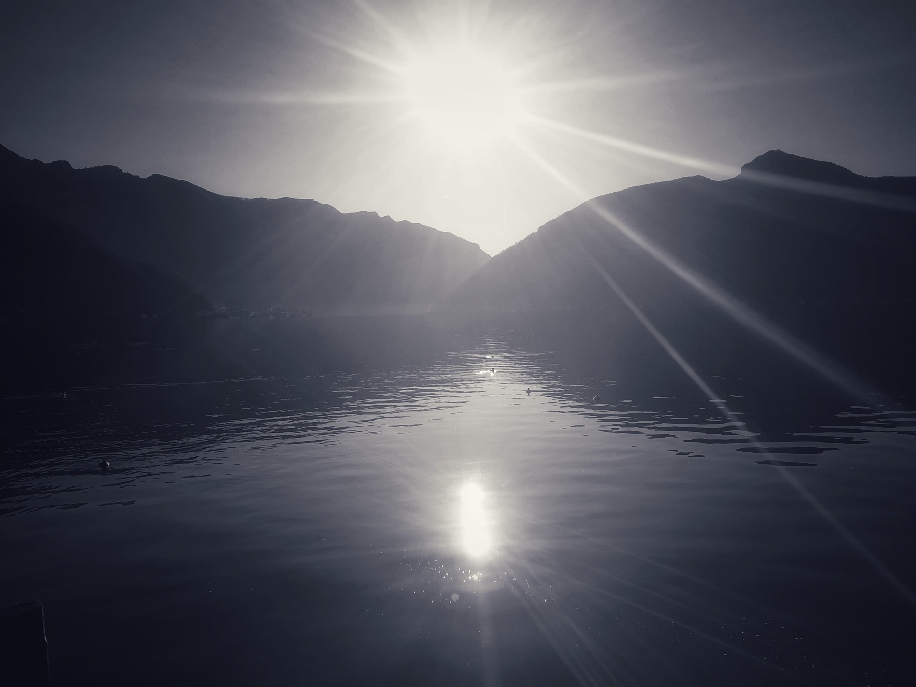 A serene landscape features a bright sun above a calm lake surrounded by silhouetted mountains.