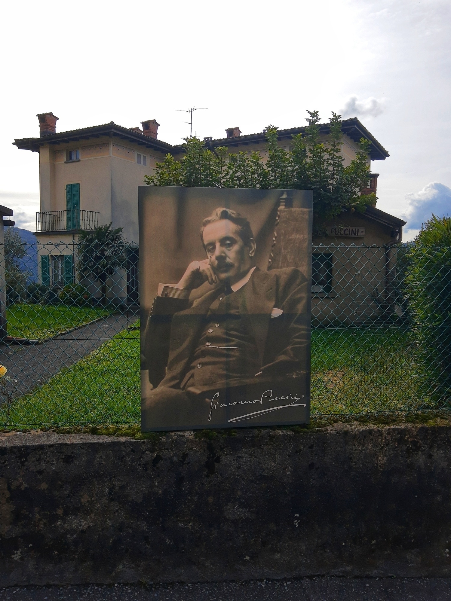 A large photograph of a man sitting thoughtfully is displayed in front of a house surrounded by a garden and fence.