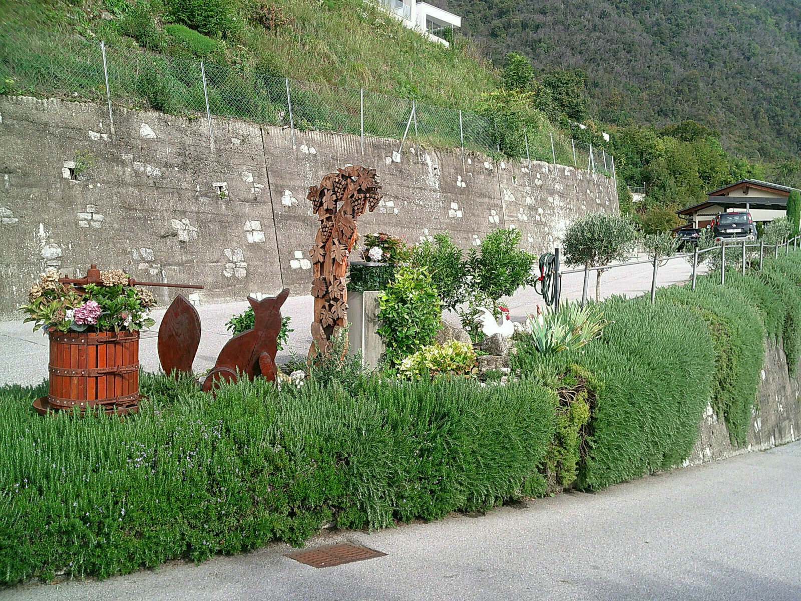 Auto-generated description: A garden by a roadside features shrubs, metal sculptures of animals, and a wooden planter with flowers.