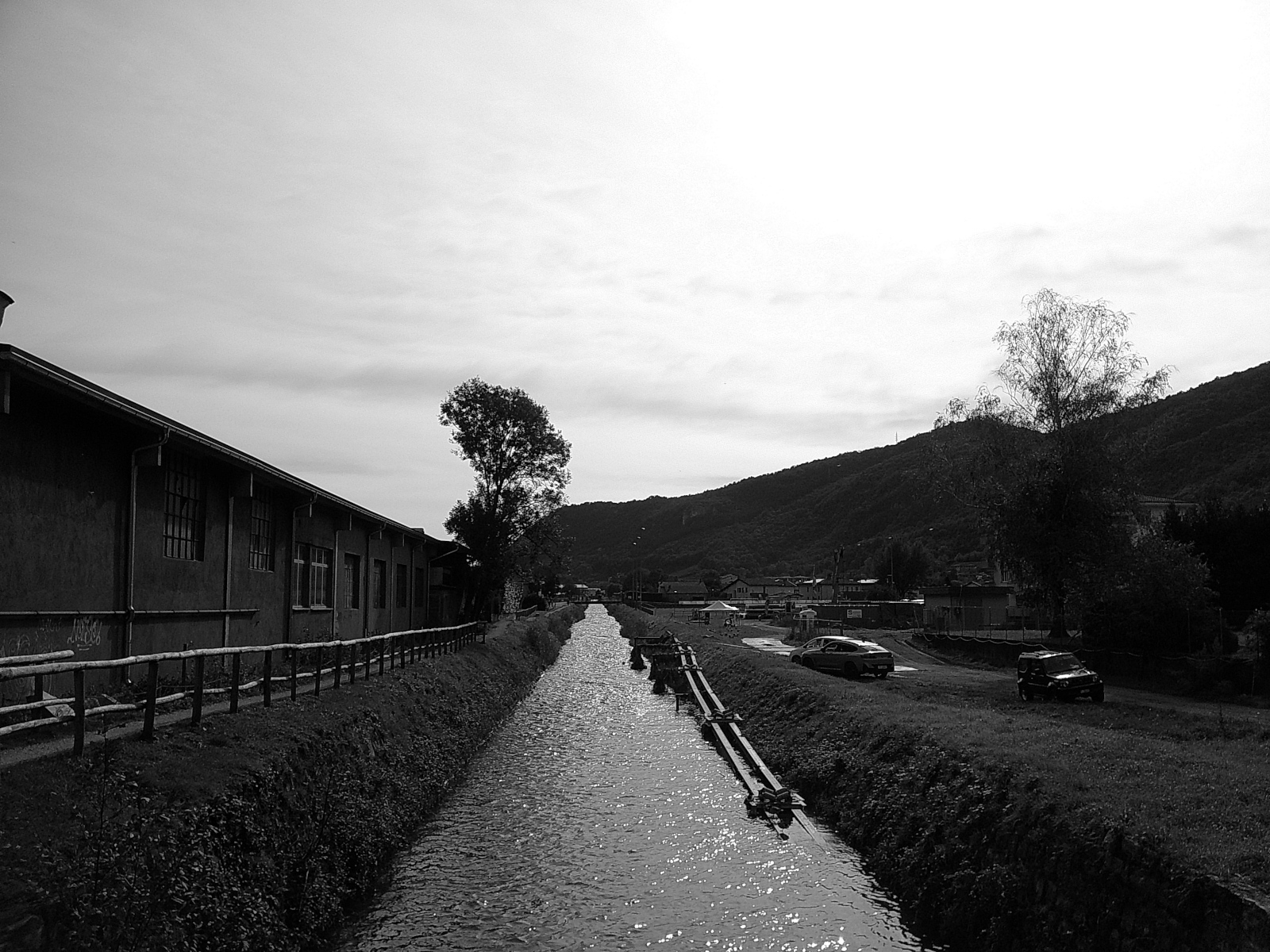 Auto-generated description: A narrow canal runs alongside a building and road with cars, set against a backdrop of hills and a large tree under a partly cloudy sky.