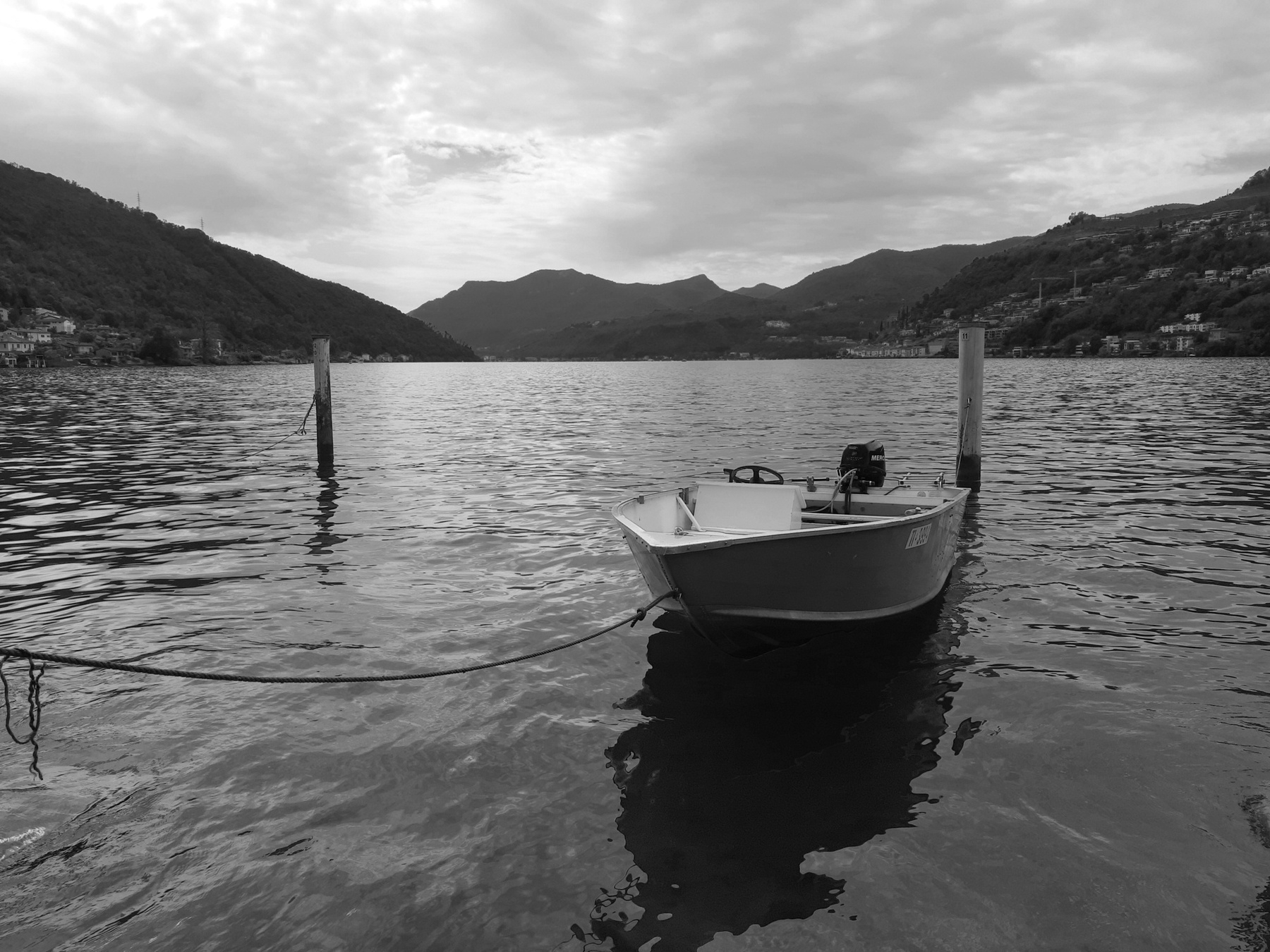 Panorama in scala di grigio con barchetta attraccata sul lago di Lugano e al di sopra delle nuvole