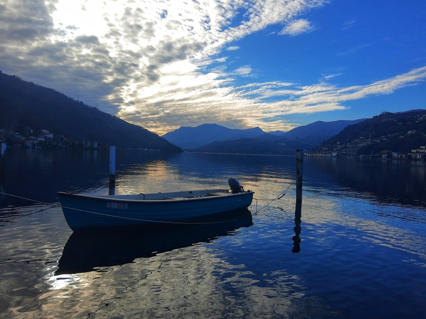 Auto-generated description: A small boat is tied to a post on a calm, reflective lake surrounded by mountains under a partly cloudy sky.