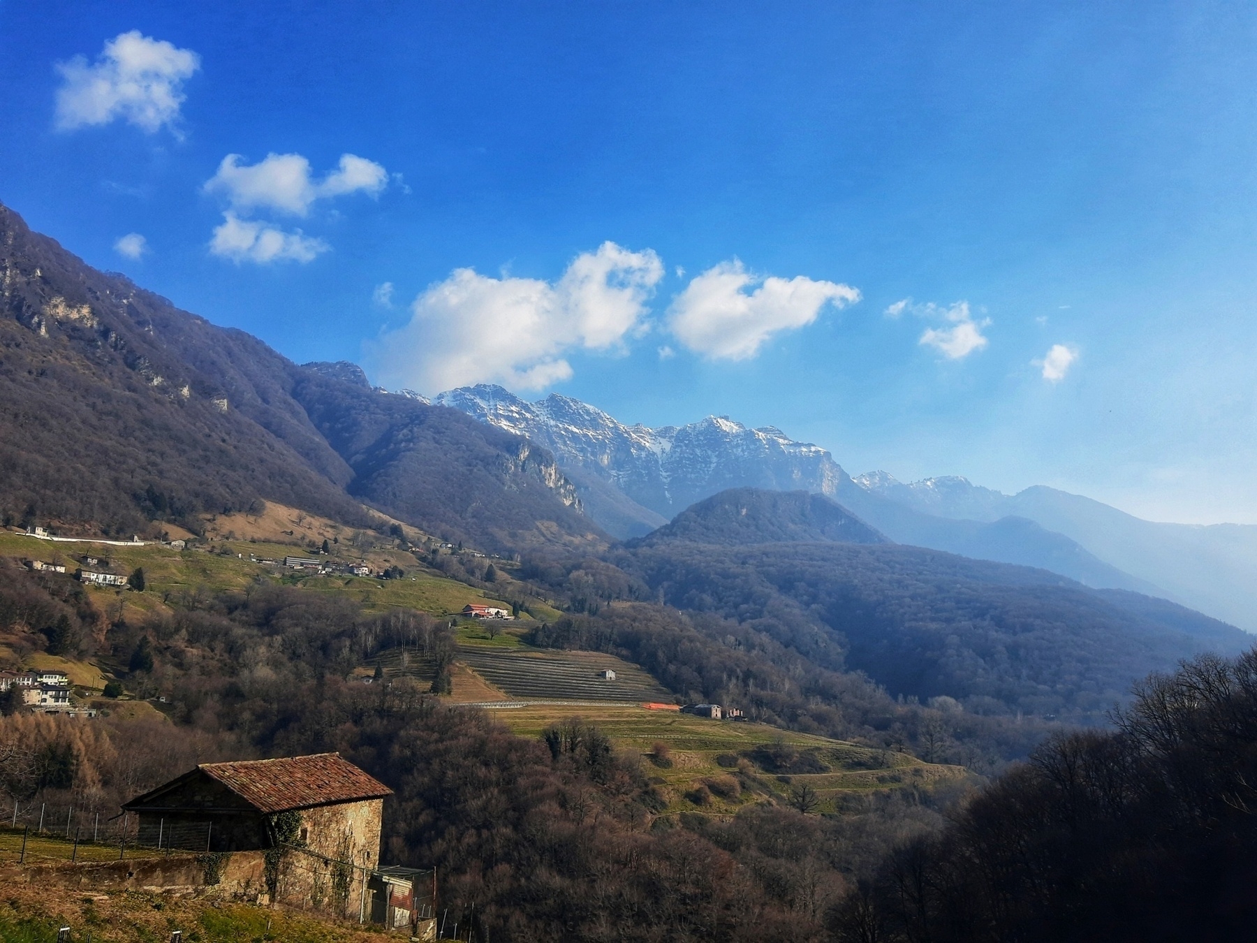 Auto-generated description: A scenic landscape features a rustic building in the foreground, rolling hills, and snow-capped mountains under a clear blue sky.