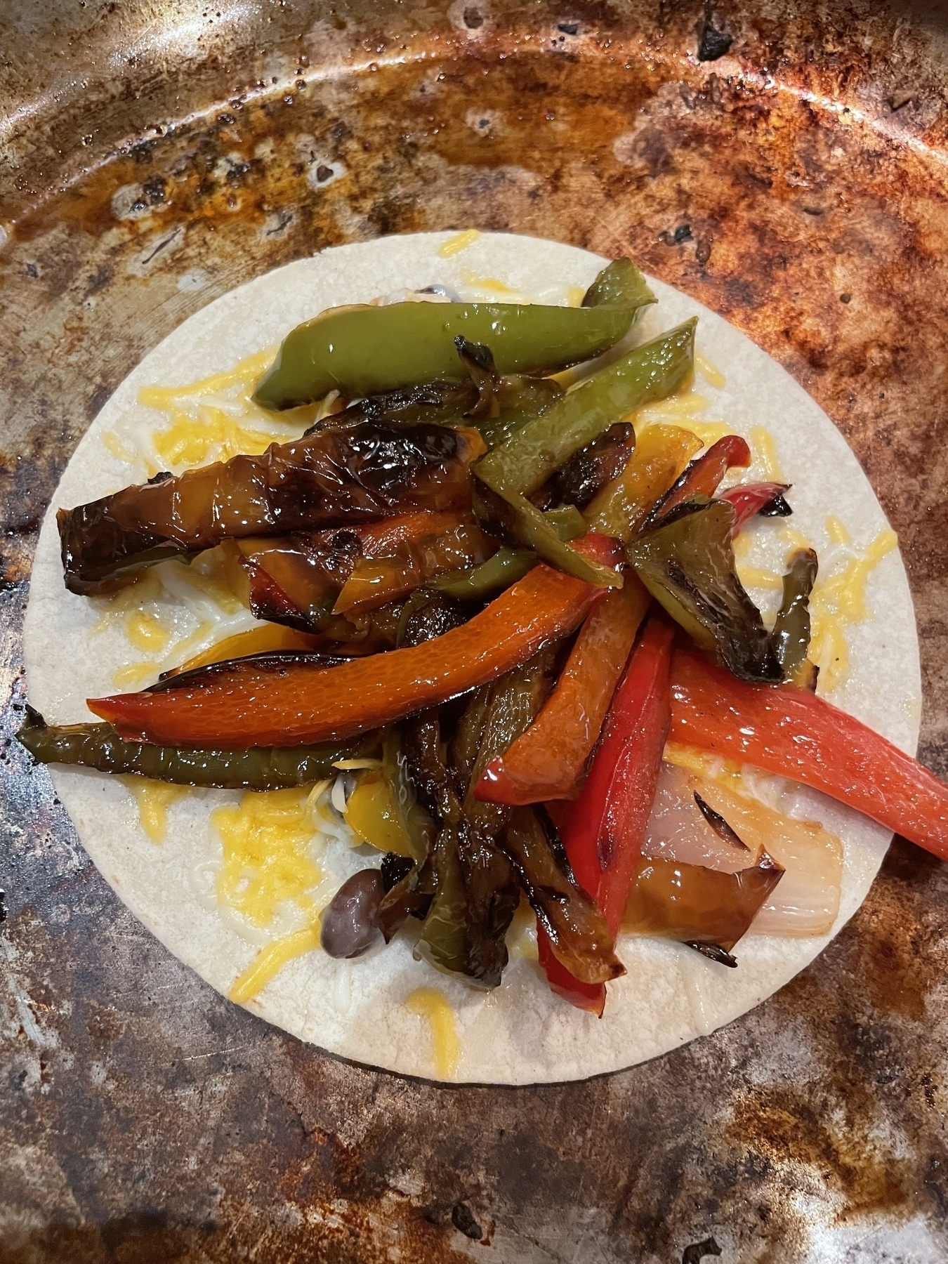 Griddled peppers and onions on some melted shredded cheese, on top of a small tortilla, cooking in a pan. More cheese and a second tortilla will be placed on top. 