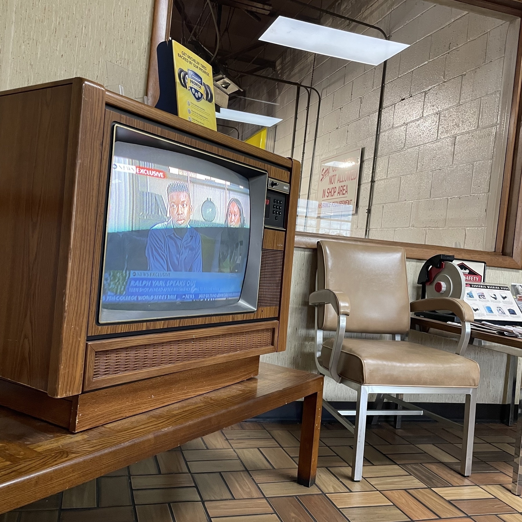 A brown wood-grain cabinet style CRT television showing live news broadcast. The TV sits on a wooden coffee table, next to a metal and vinyl upholstered waiting room chair. 