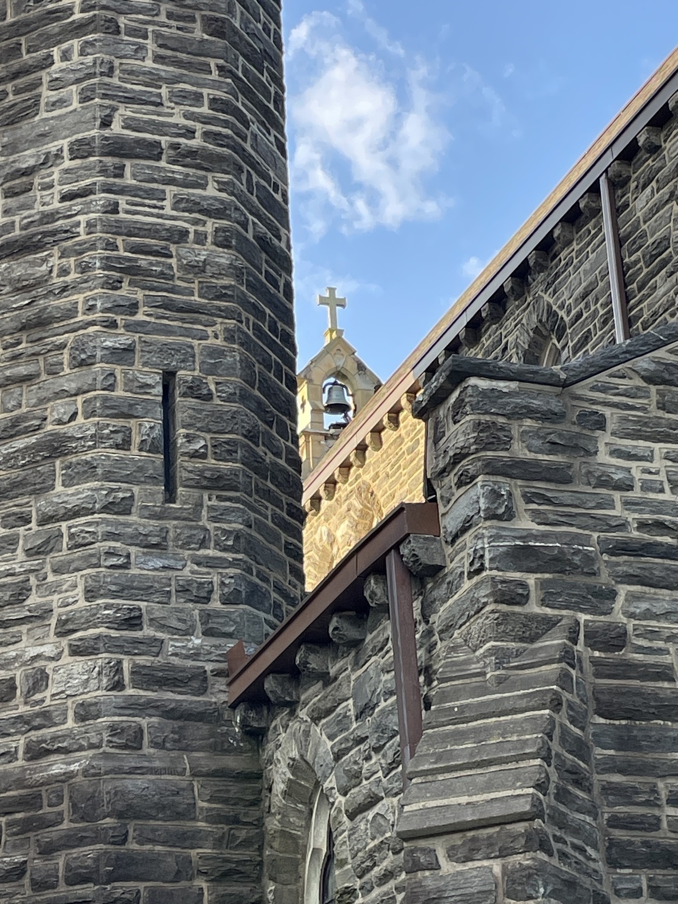 Photo of the outside of a gothic stone church, where a stone tower connected to some interestingly angled walls is in the foreground and a brighter lit bell tower with cross on top is in the middle background. &10;