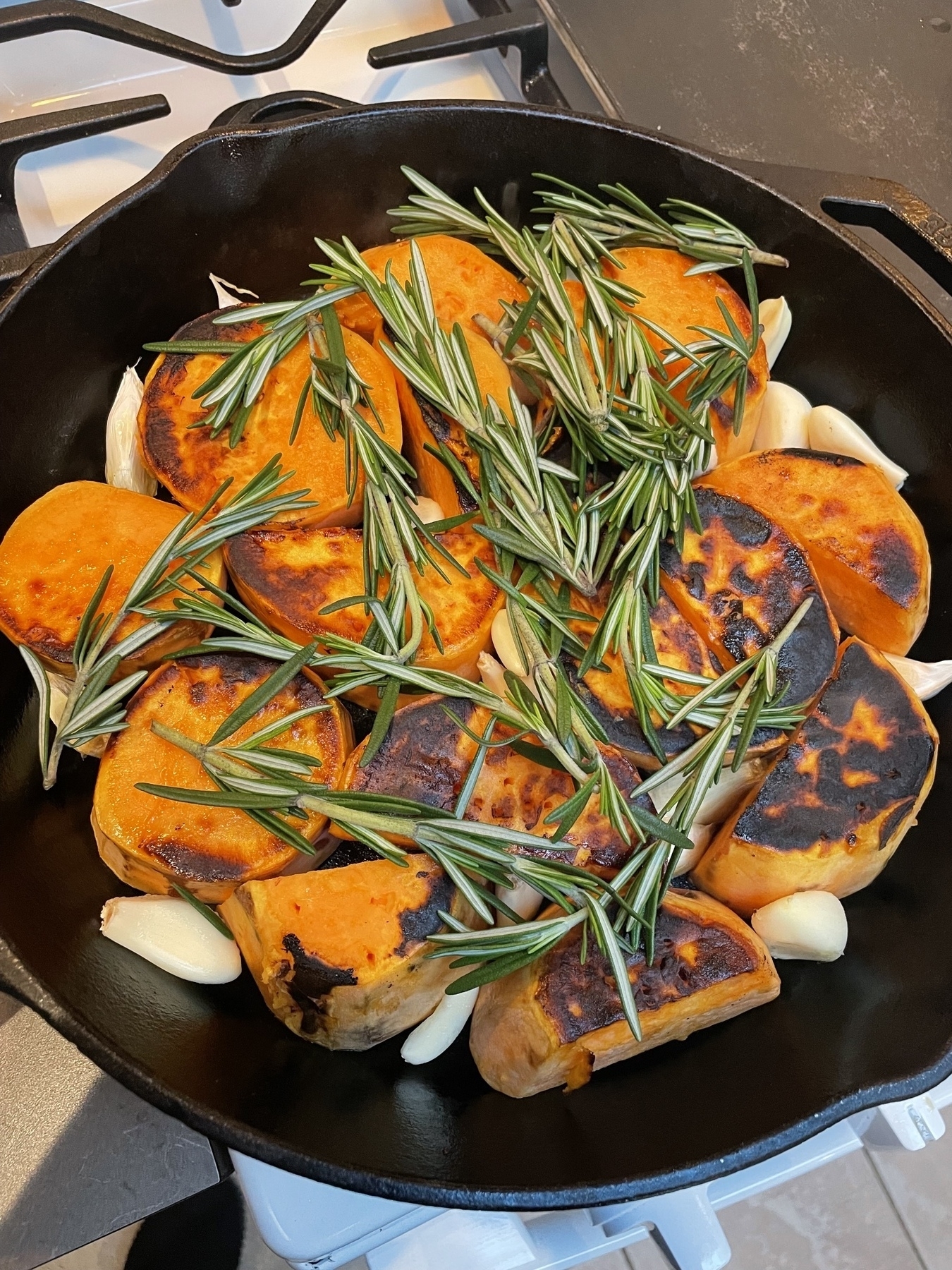 Beautifully orange sweet potato chunks placed in a black iron skillet and already seared & caramelized some on one side (some dark blistering on the potato surfaces) with fresh garlic cloves sprinkled around the potatoes, and a handful of rosemary sprigs draped over the top 