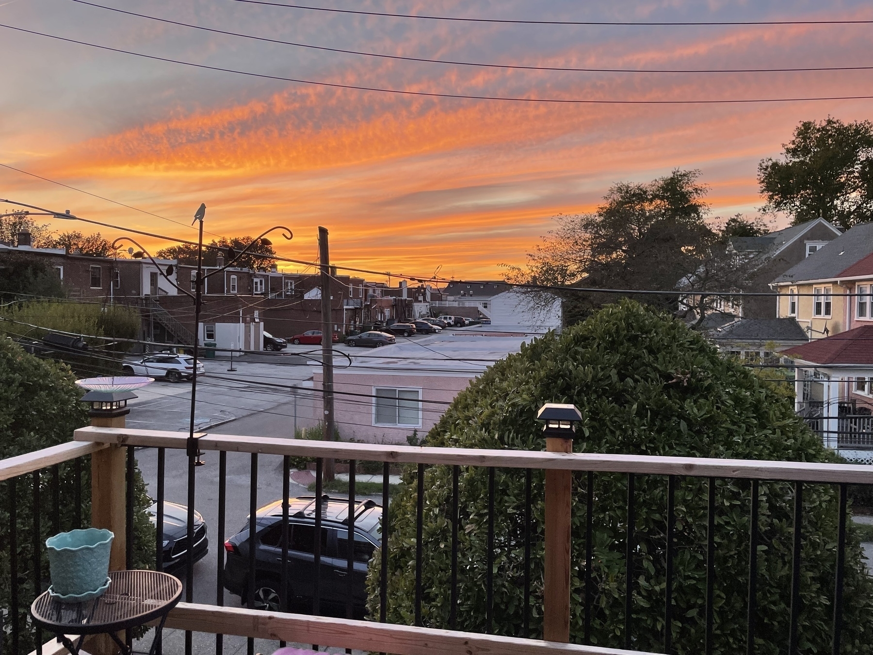 Twilight sky — with skeins of long clouds hanging in the air like a giant fuzzy blanket of blue, yellow-orange — over a balcony railing across an urban street with some buildings in the distance. 