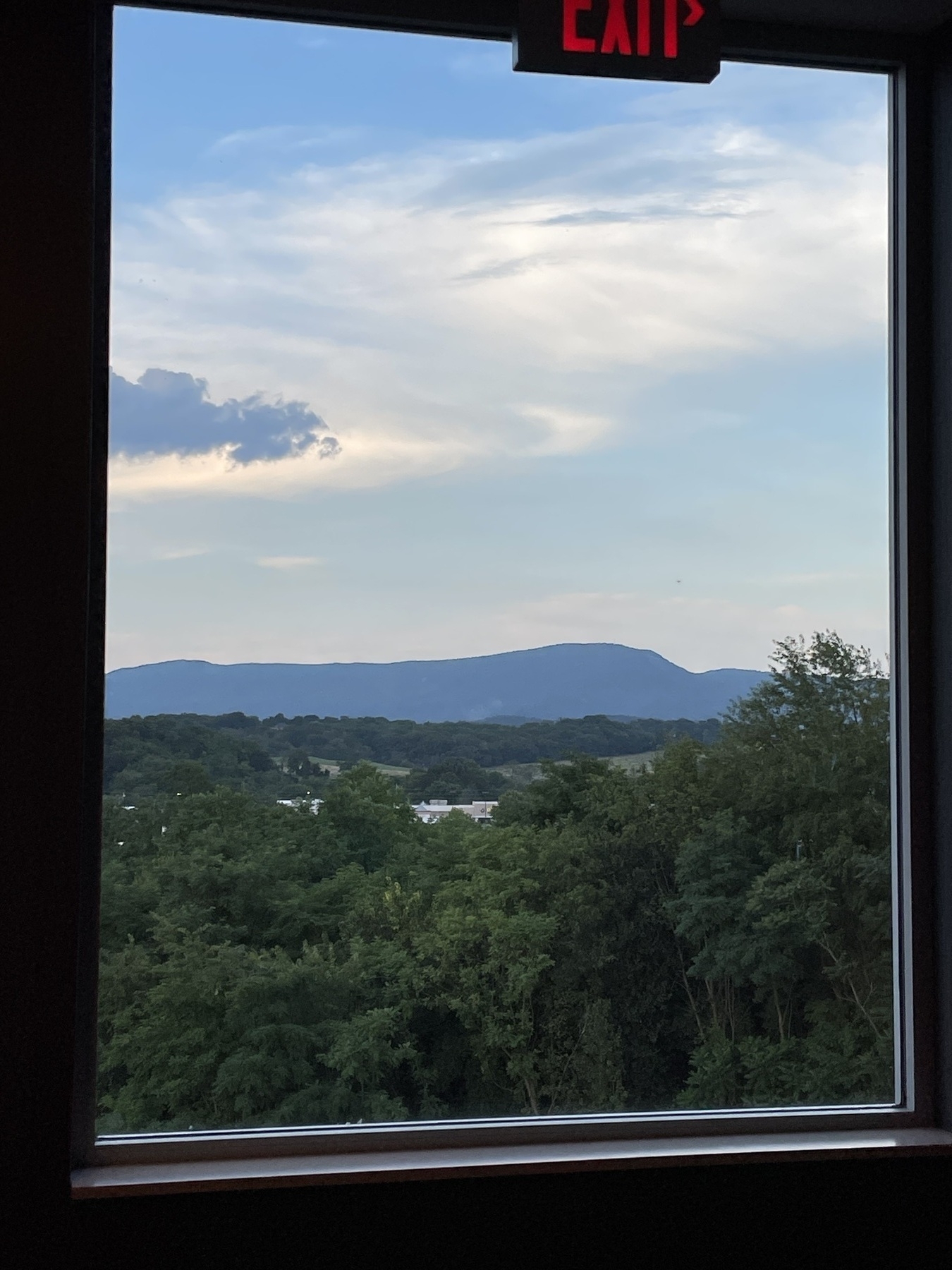 Photo showing landscape through a broad single pane window frame from our hotel. The landscape is beautiful with swirling bits of cloud in a blue sky, allow range of hills in the distance, and green forest in the foreground. 