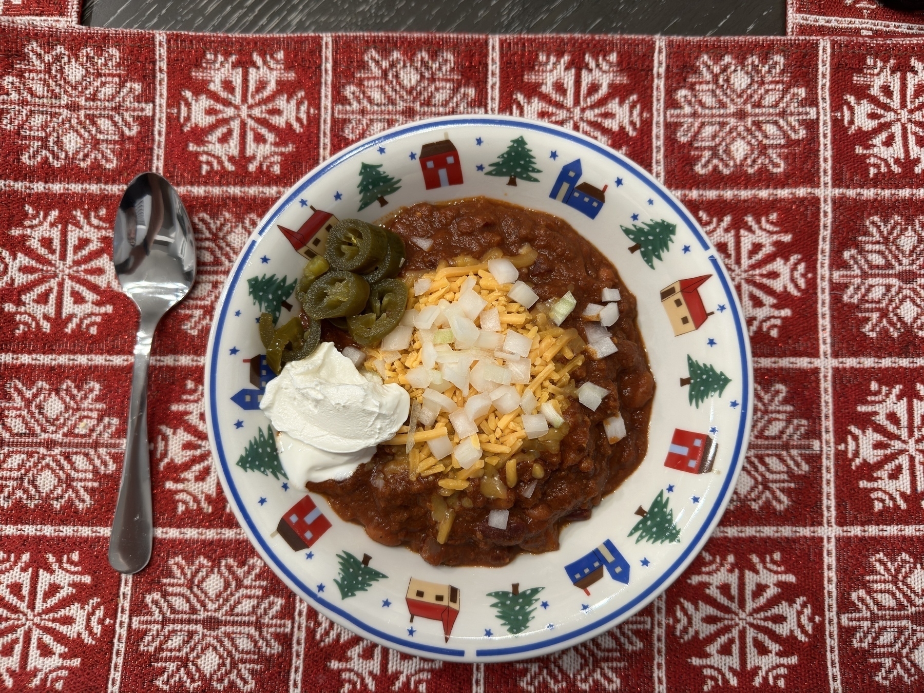 A bowl of homemade chili with cheese, a dollop of sour cream and jalapenos on a holiday placemat. A spoon sits to the left of the bowl.