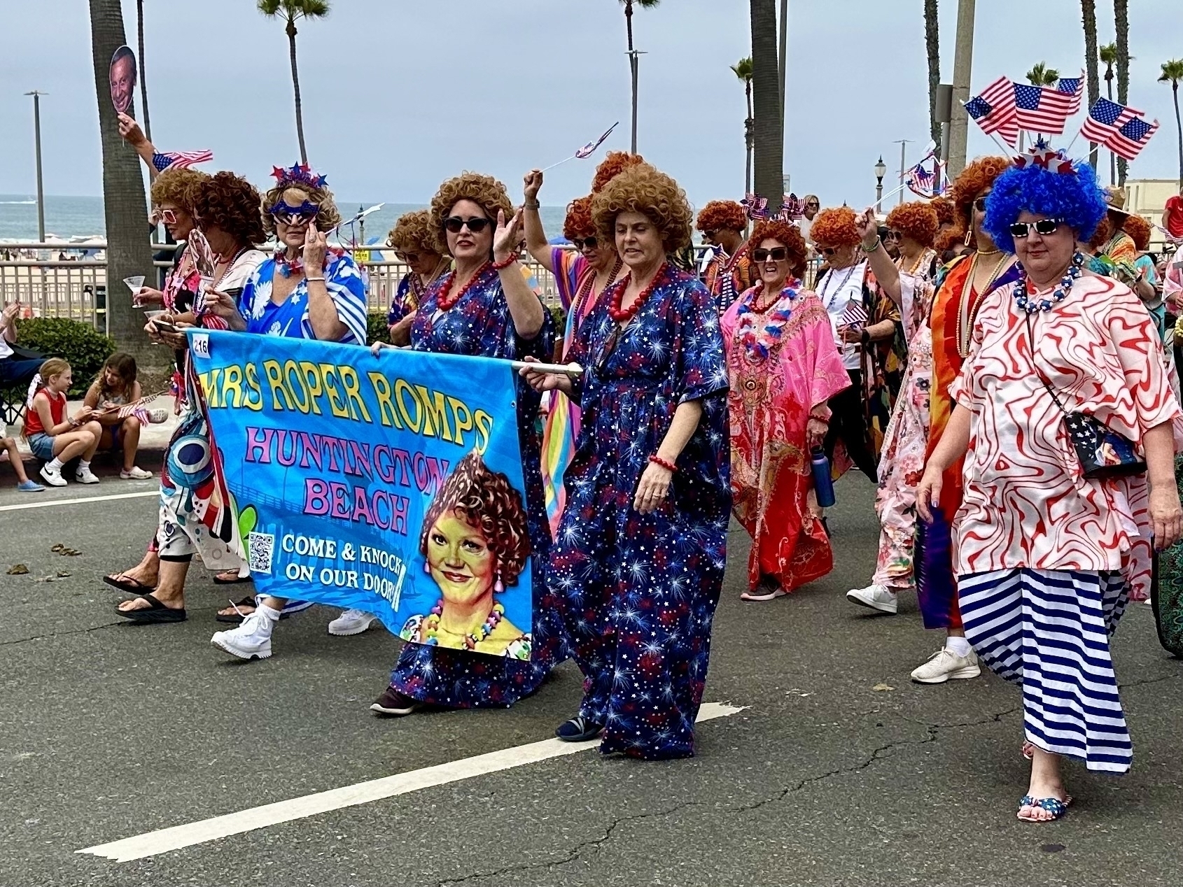 A group of people dressed in vibrant, 1970s-style costumes and wigs are marching in a parade, holding a banner that reads "Mrs. Roper Romps Huntington Beach."