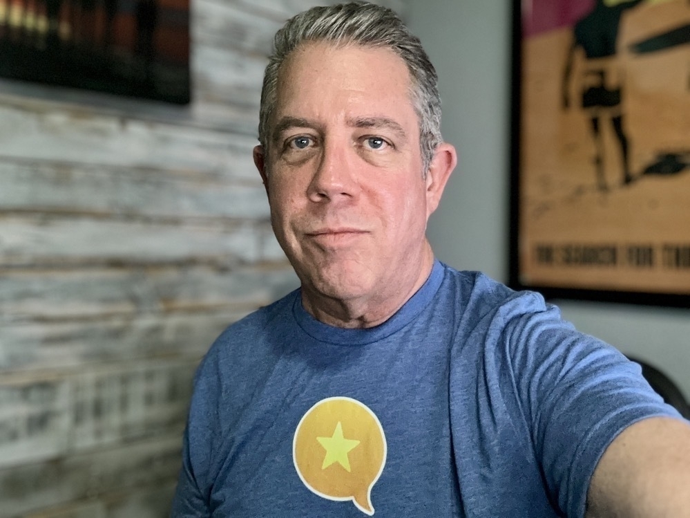 A person wearing a blue shirt with a yellow Micro.blog logo design stands in a room with posters and wood paneling on the walls.