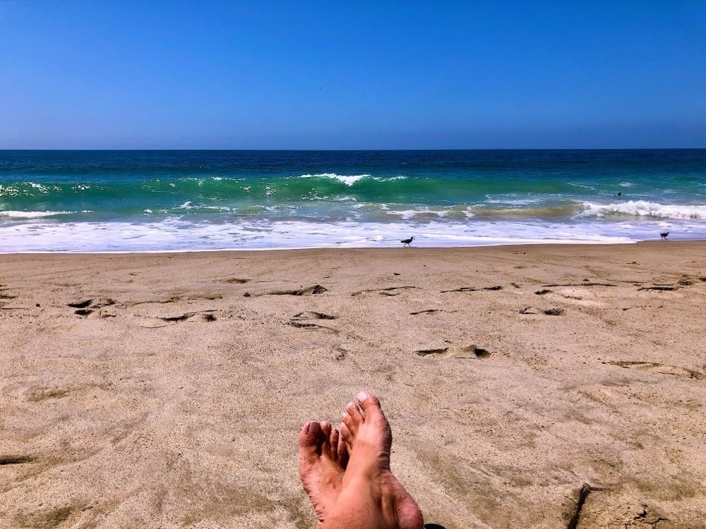Person's bare feet on sandy beach with ocean waves in background.