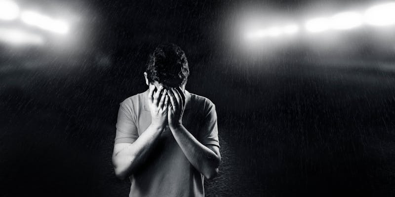 Auto-generated description: A person standing in a dimly lit scene covers their face with their hands, surrounded by falling rain.