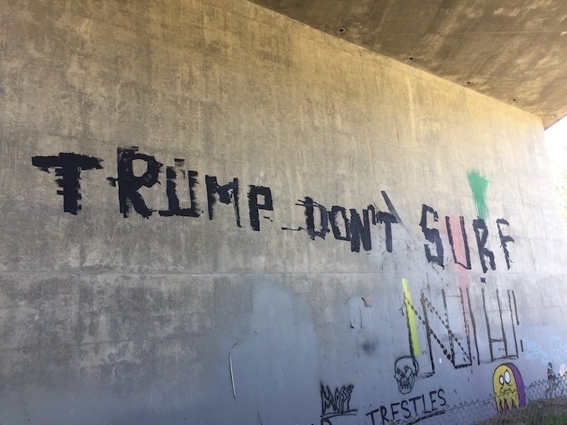 Graffiti on a concrete wall reads 'TRUMP DON'T SURF' in black letters along with other smaller drawings and writings nearby.