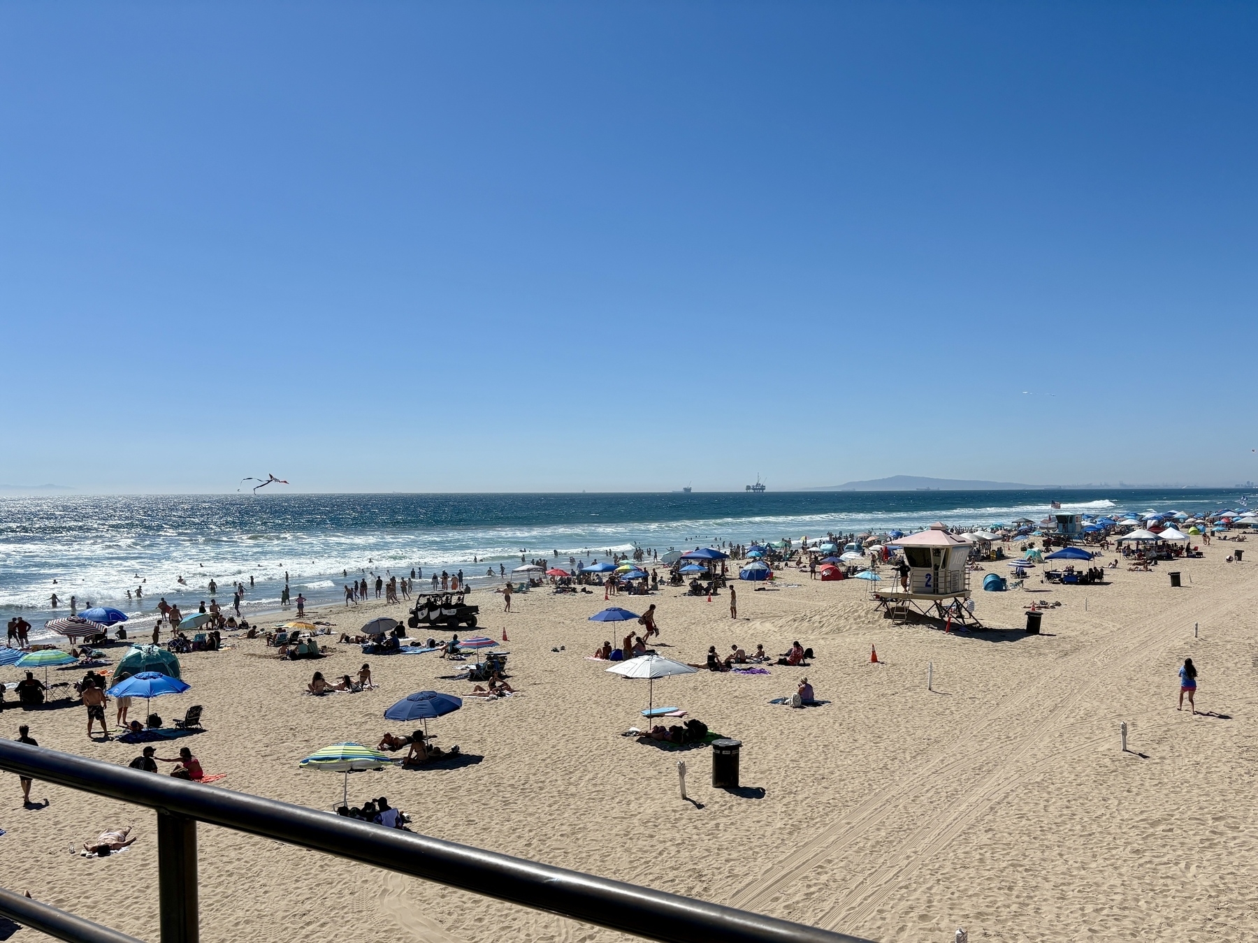 A crowded beach scene features numerous people enjoying the sand and sea under a clear blue sky, with colorful umbrellas scattered across the shore.