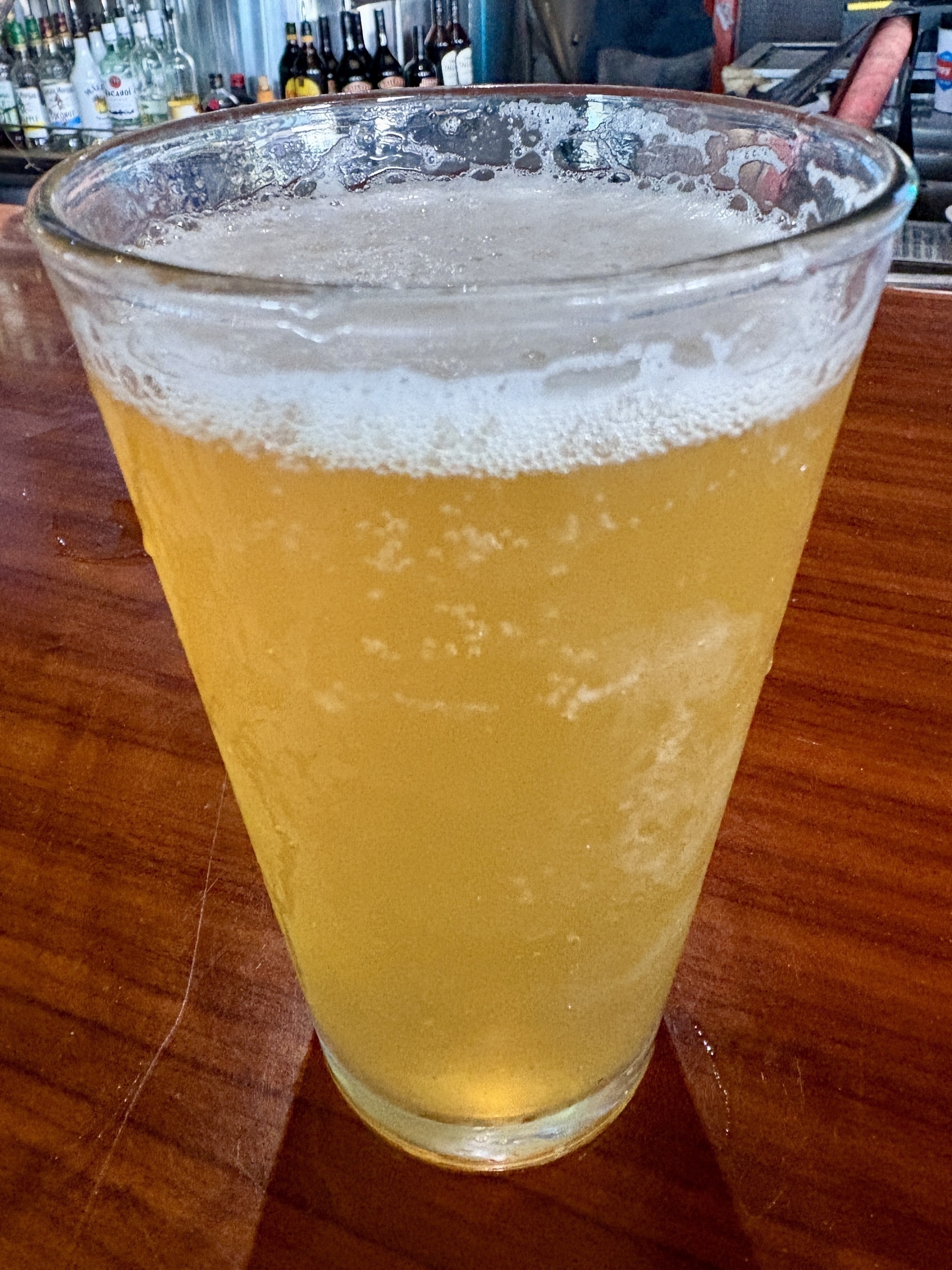 A frothy glass of beer sits on a wooden bar counter.