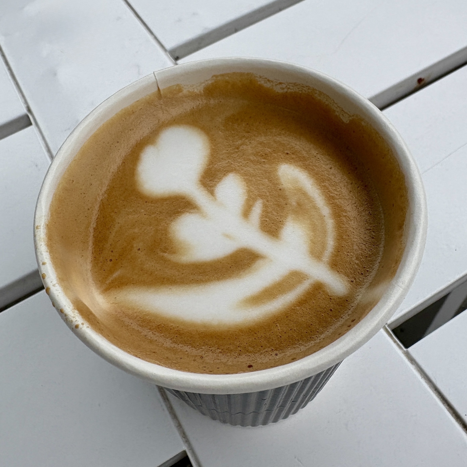 A latte in a paper cup features beautiful flower-shaped latte art on a white table.