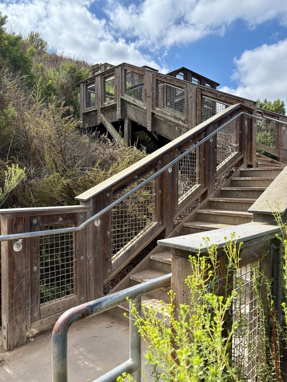 A wooden staircase with metal railings leads up to a scenic overlook surrounded by greenery and blue skies.