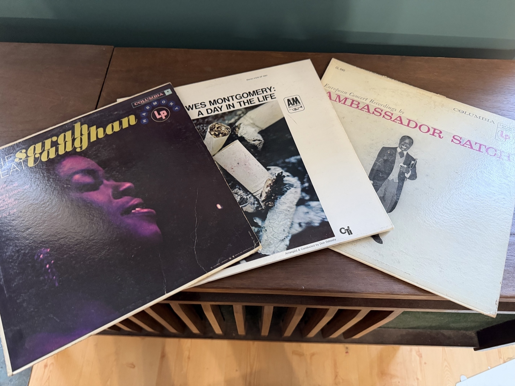 3 vinyl record albums are displayed on a wooden surface, &10;The Great Sarah Vaughan HL7158 Columbia records (title in yellow font and purple hued pic of Sarah in profile)&10;Wes Montgomery a day in the life - text in black on white album cover with black and whit photo of cigarette butts.&10;Ambassador Satch - white album with title in Red font with black and white full body pic of Louis Armstrong smiling holding a white kerchief up to his chest in his left hand and his trumpet in the crook of his left arm.  His right holds a suitcase at his side.