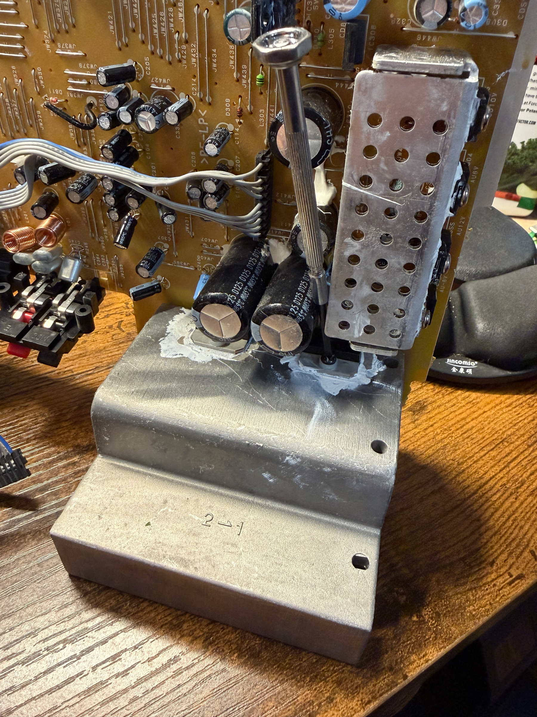 A circuit board with a soldering iron and metal heatsink attached is set on a wooden surface.