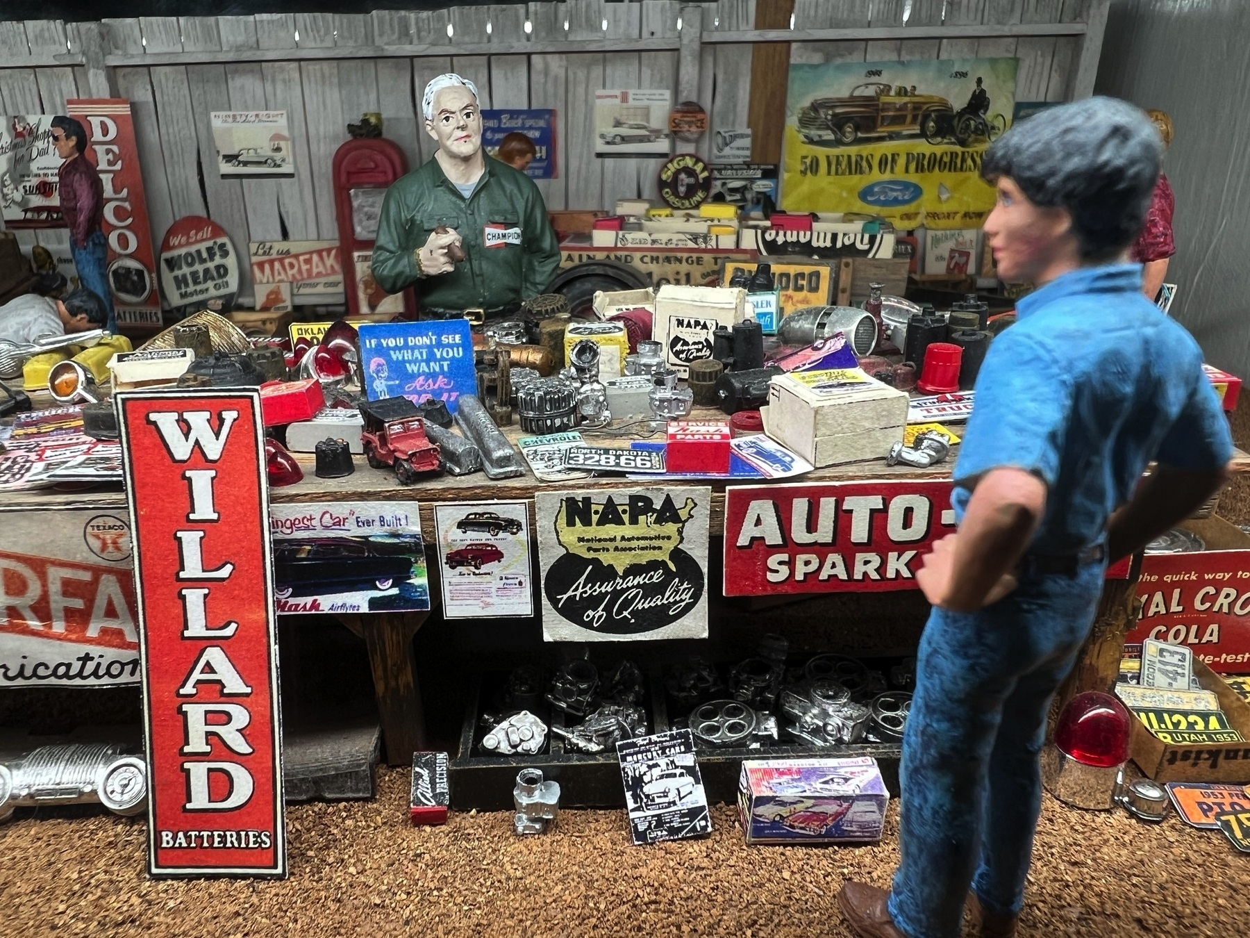 Minitature set up with two people are in a garage-like setting filled with vintage automotive signs and various car parts.