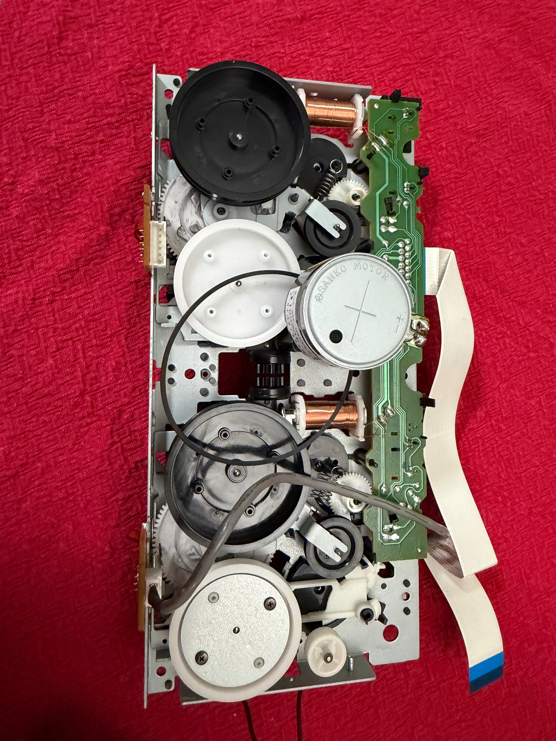 A mechanical assembly with gears, spools, and a circuit board is placed on a red textured surface.