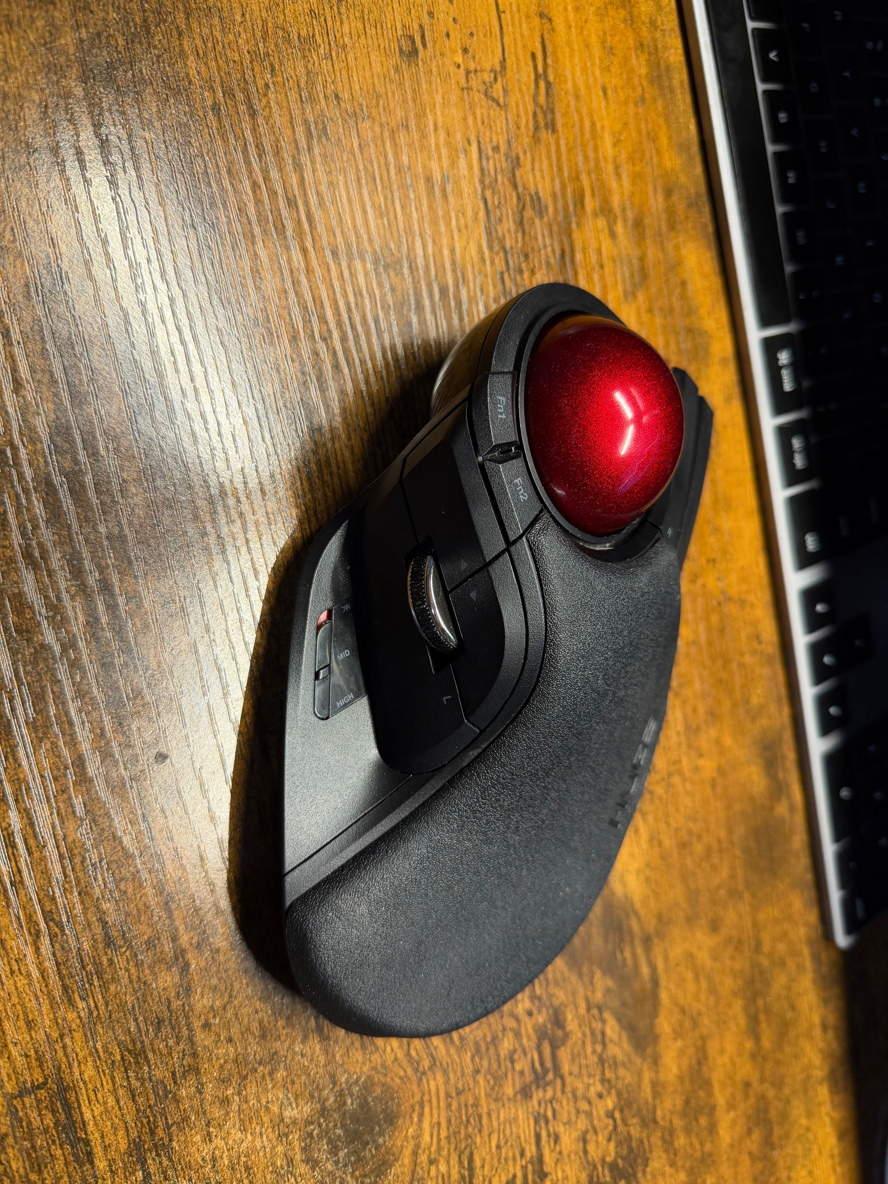 A red ball wireless ergonomic mouse is placed on a wooden surface next to a keyboard.
