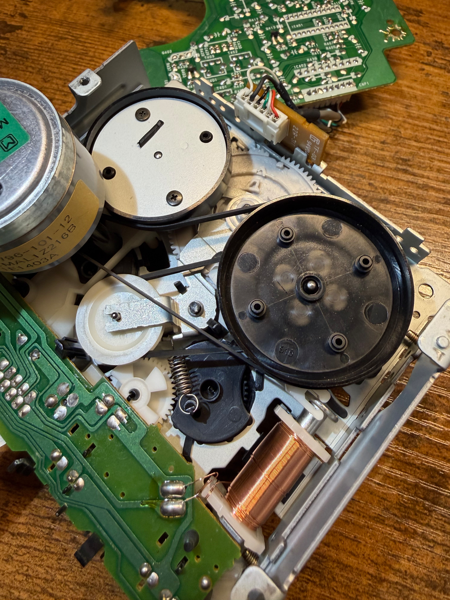 A disassembled tape mechanism showing various gears, circuits, and components on a wooden surface.  Clearly visible motor in upper left and new belts around gears and fly wheel.