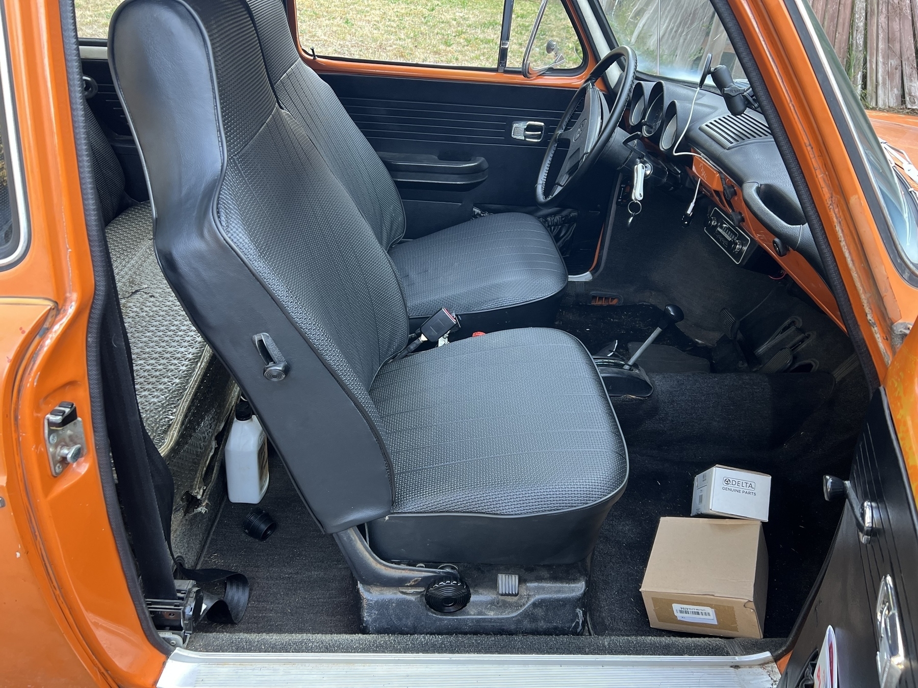 An interior view of a vw type 3 car shows black seat covers, a simple dashboard, and two boxes on the floor.