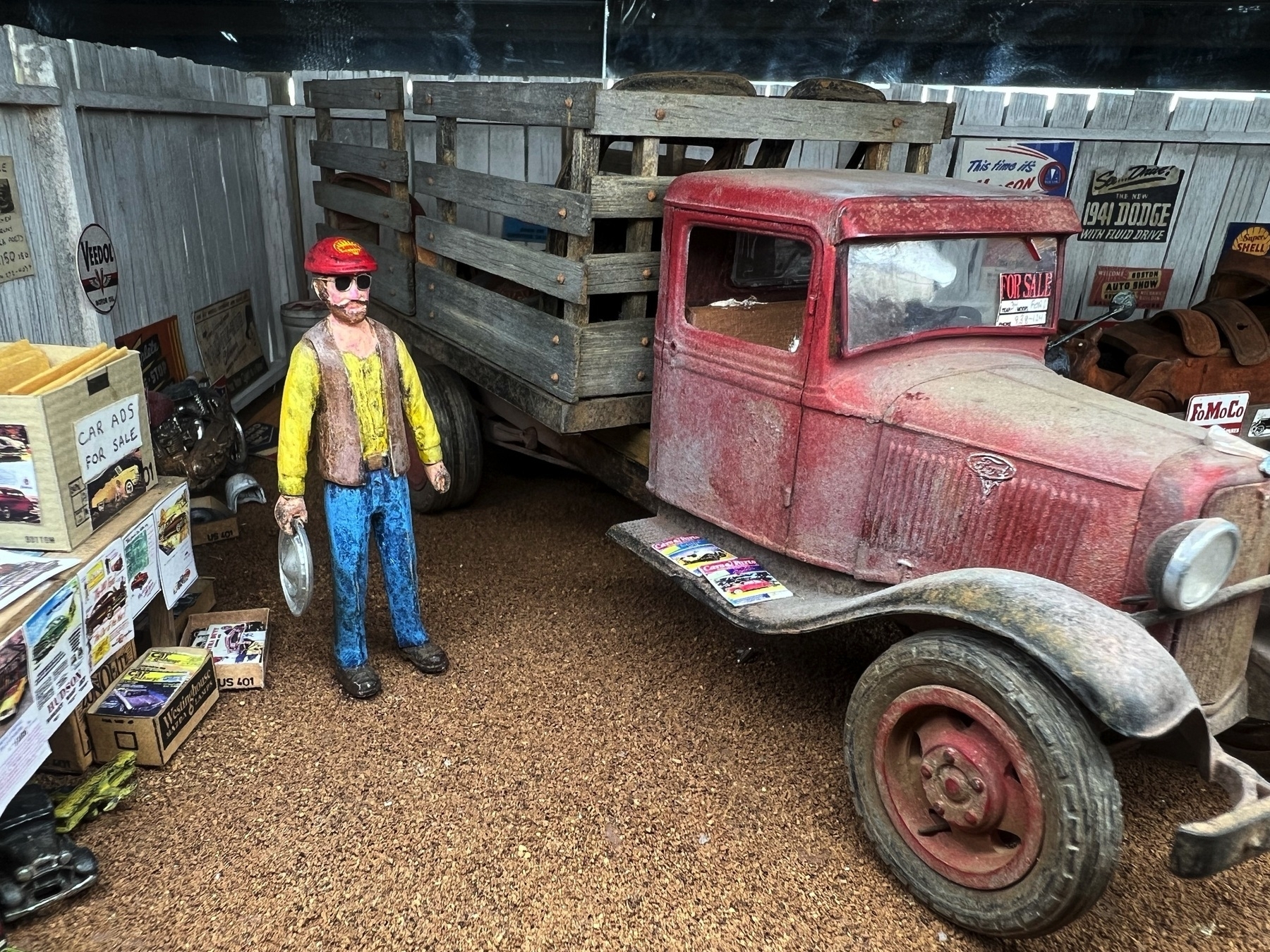 A miniture vintage red truck is parked inside a shed, accompanied by a colorful statue of a man holding a pan, surrounded by boxes and signs.