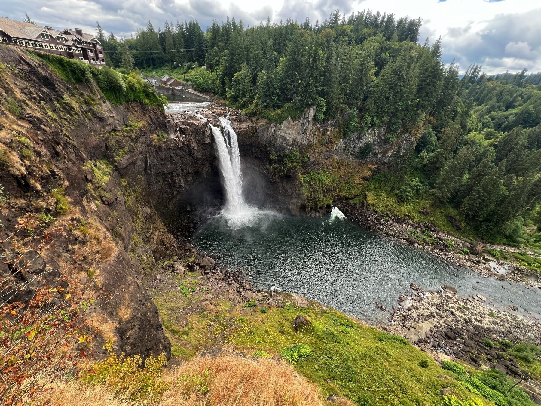 A waterfall cascades into a rocky pool surrounded by lush green trees, with a building located at the top left of the scene.