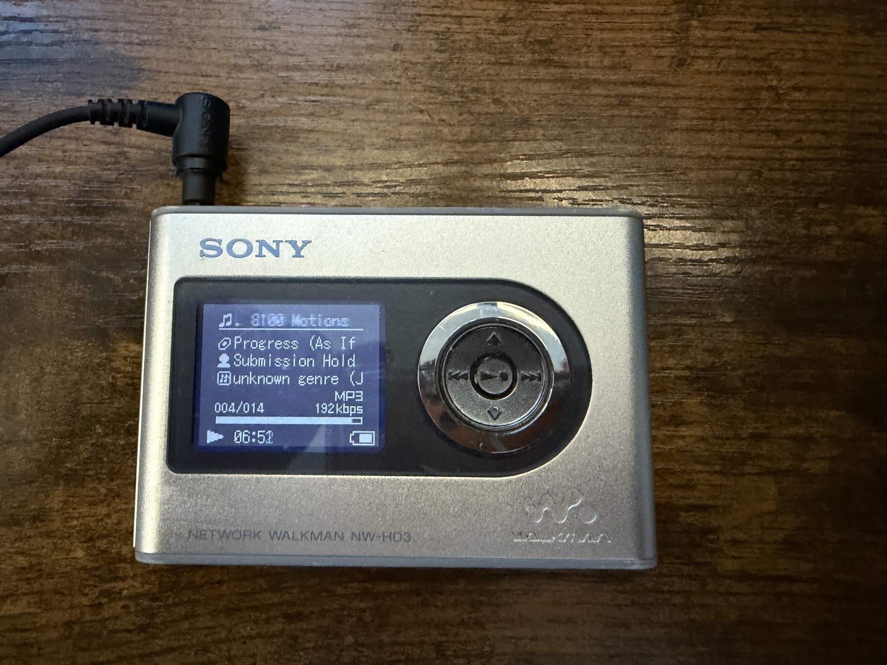 A silver Sony Network Walkman NW-HD3 displaying a music track on its screen is resting on a wooden surface.