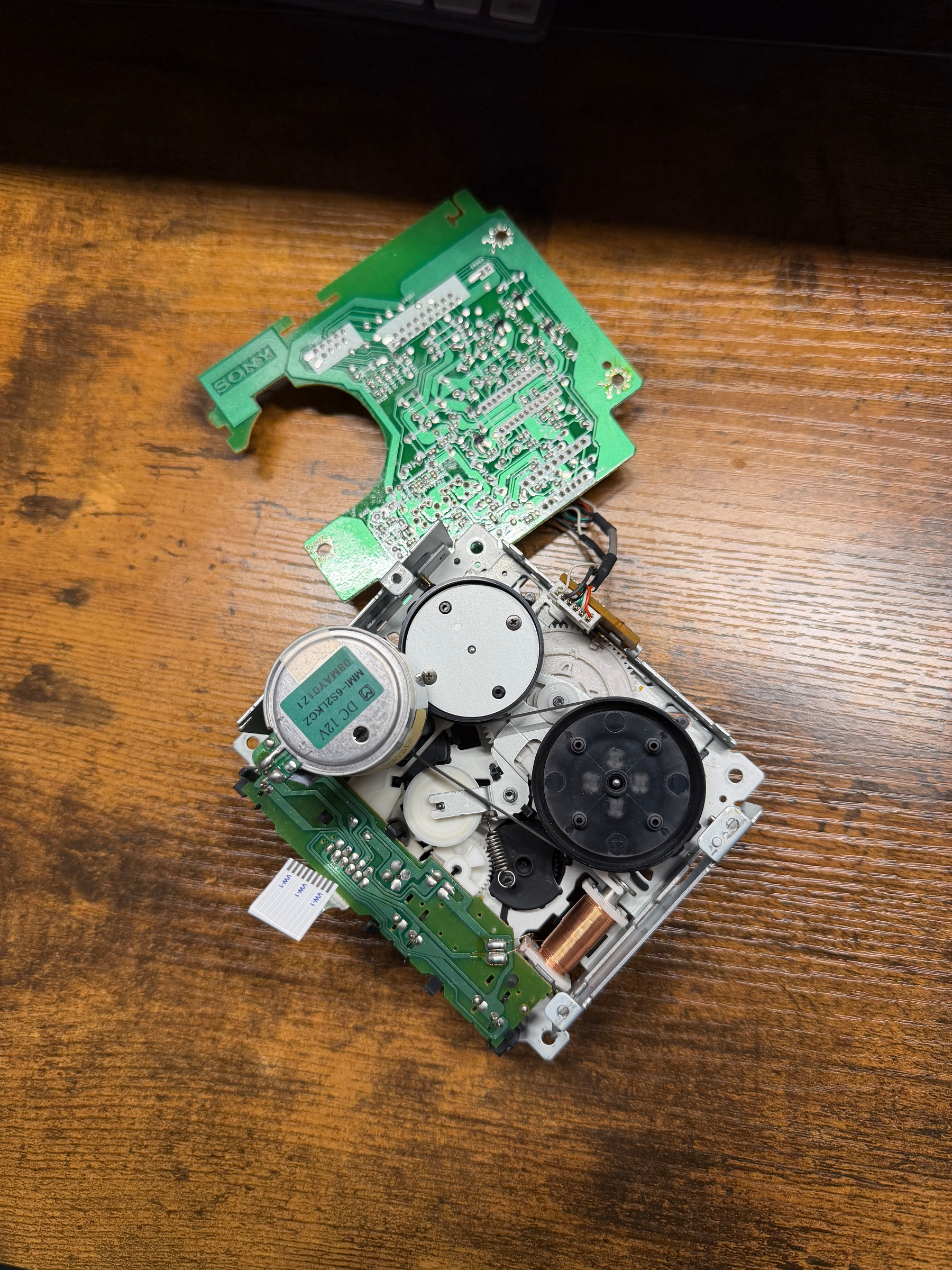 A disassembled tape mechanism showing circuit boards, gears, and mechanical components rests on a wooden surface.