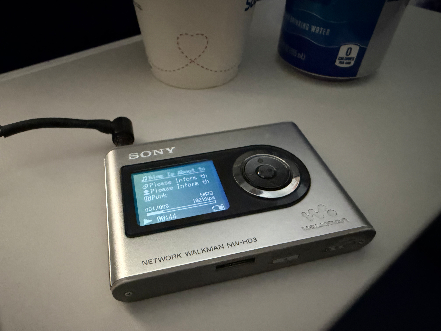 A silver Sony Network Walkman NW-HD3 with an attached headphone cable is placed on an airplane seat back tray with a cup and a can next to it at top of frame.