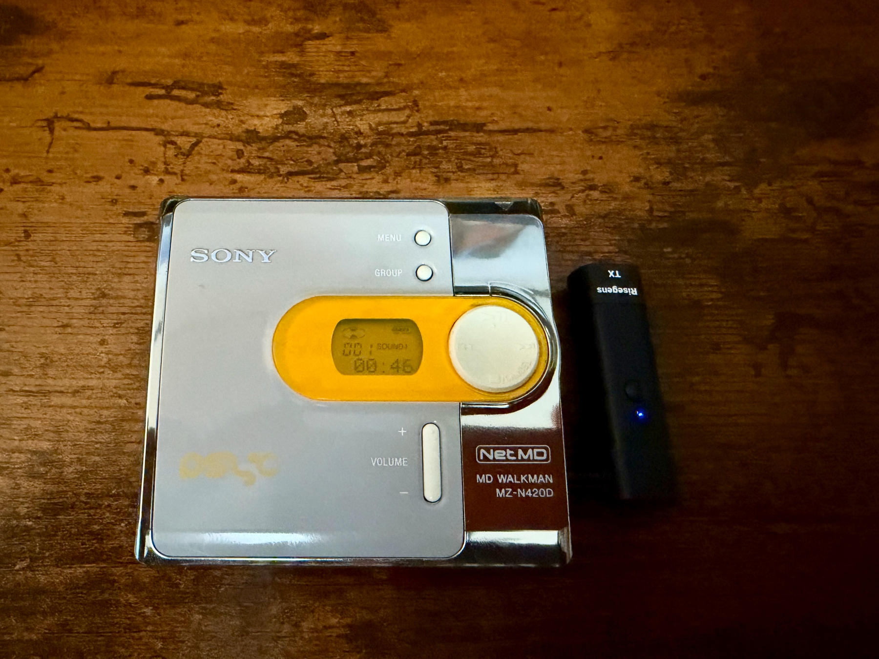 A silver Sony NetMD MD MZ-N420D Walkman with a small black bluetooth transmitter plugged into the headphone jack placed on a wooden surface.