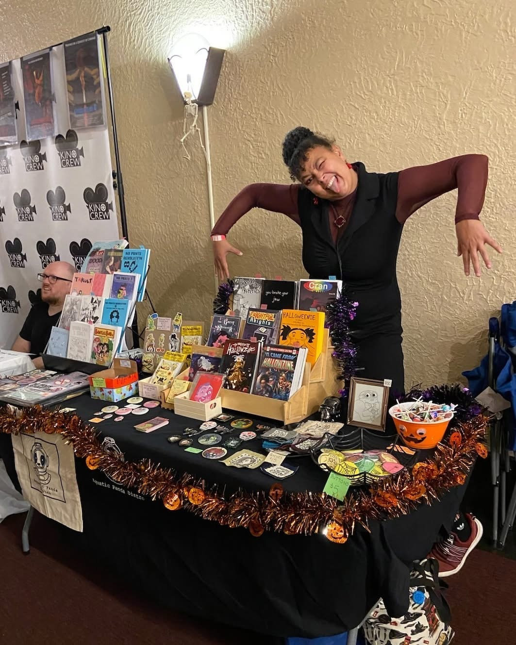 Andrea enthusiastically poses behind their table decorated with Halloween-themed items and various zines at the Aquatic Panda Distro booth.