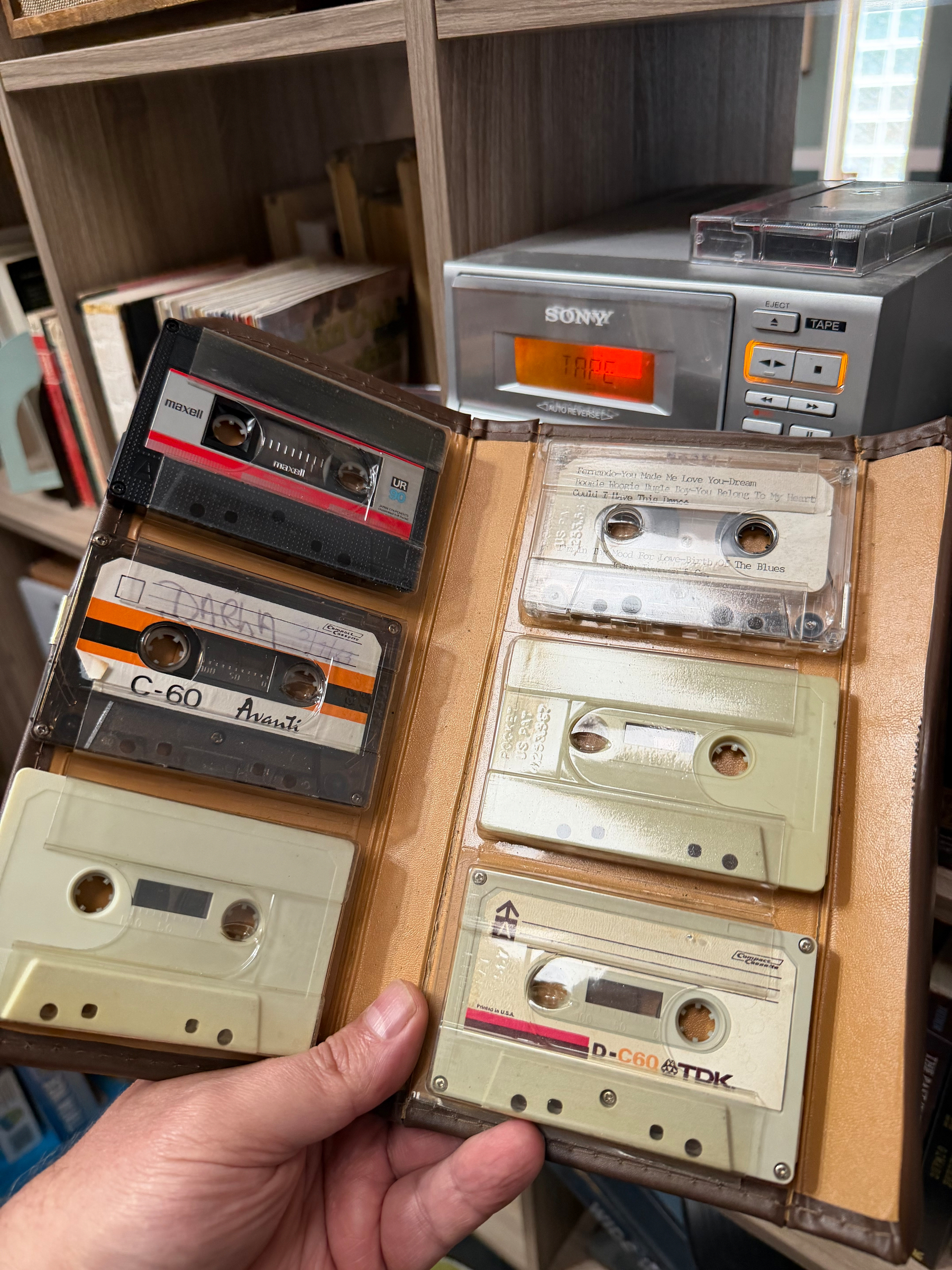 A person is holding a cassette tape organizer with several tapes inside, in front of a cassette player and bookshelves.