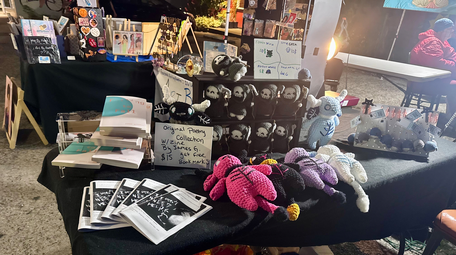A vendor table displays various handmade items including crocheted animals, a collection of poetry booklets, and other crafts arranged neatly under bright lighting.
