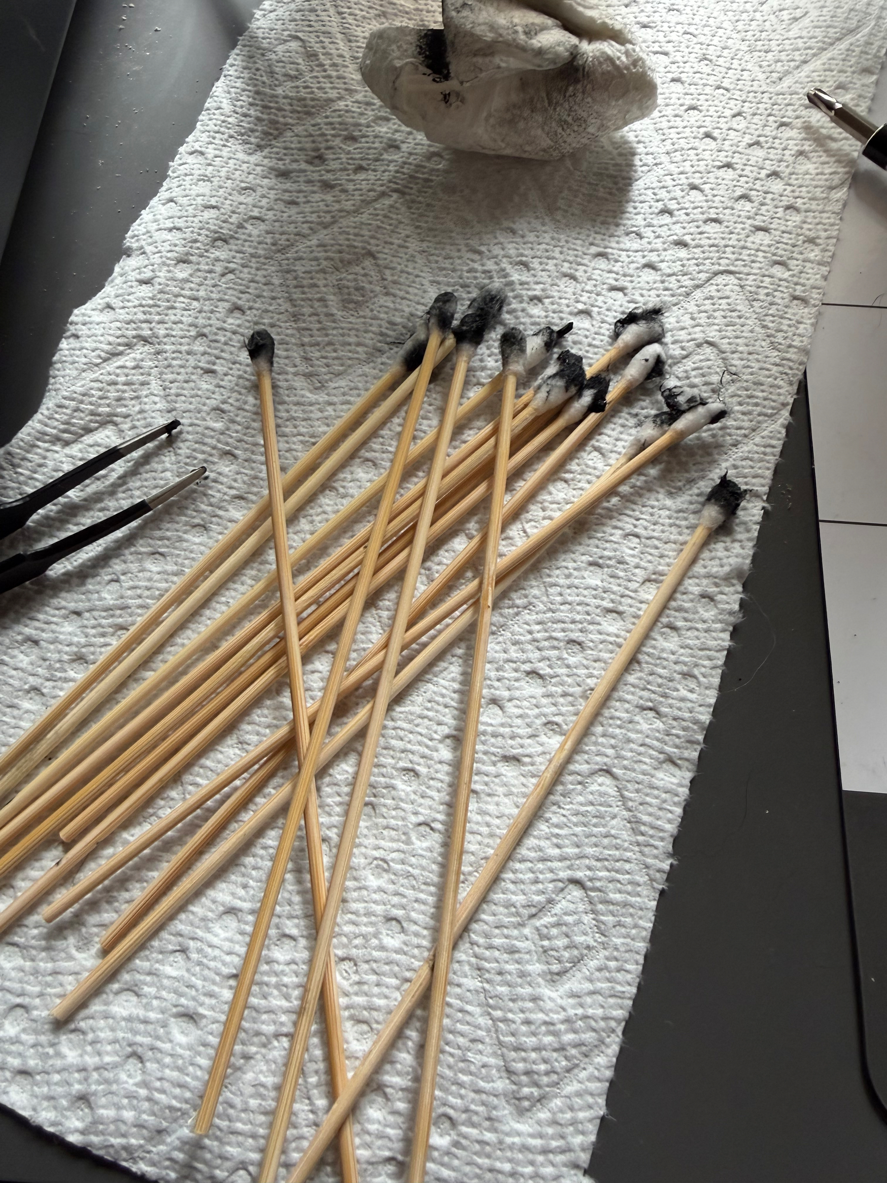 A collection of long wooden cotton swabs on their ends is placed on a paper towel next to a pair of tweezers and a crumpled paper.  Their ends are black with the goo of old rubber cassette belts.